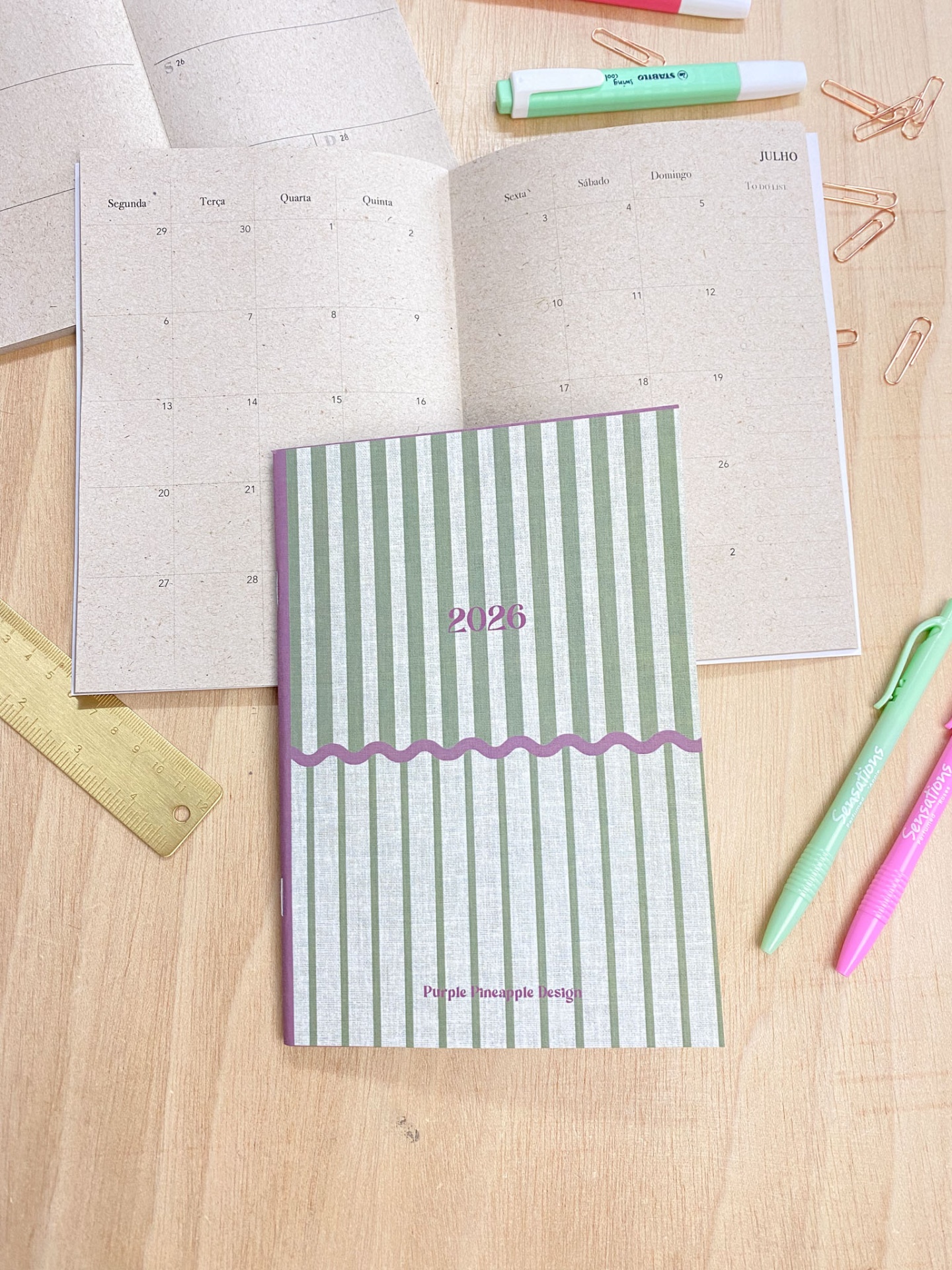 Agenda 2026 com capa verde e branca às riscas, aberta em calendário mensal com canetas e régua sobre mesa