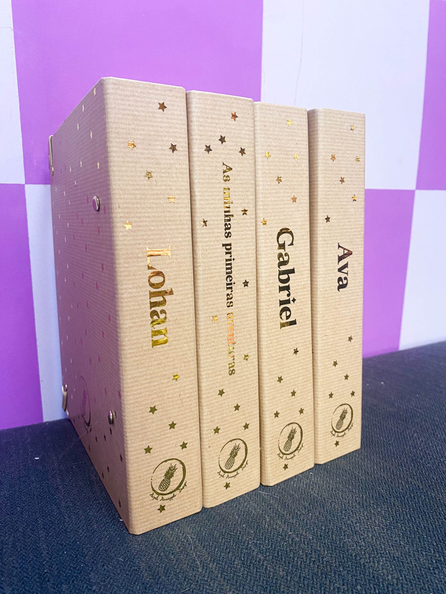 Quatro álbuns de fotografia com capas bege e estrelas douradas, contendo nomes e texto, em fundo quadriculado roxo e branco.