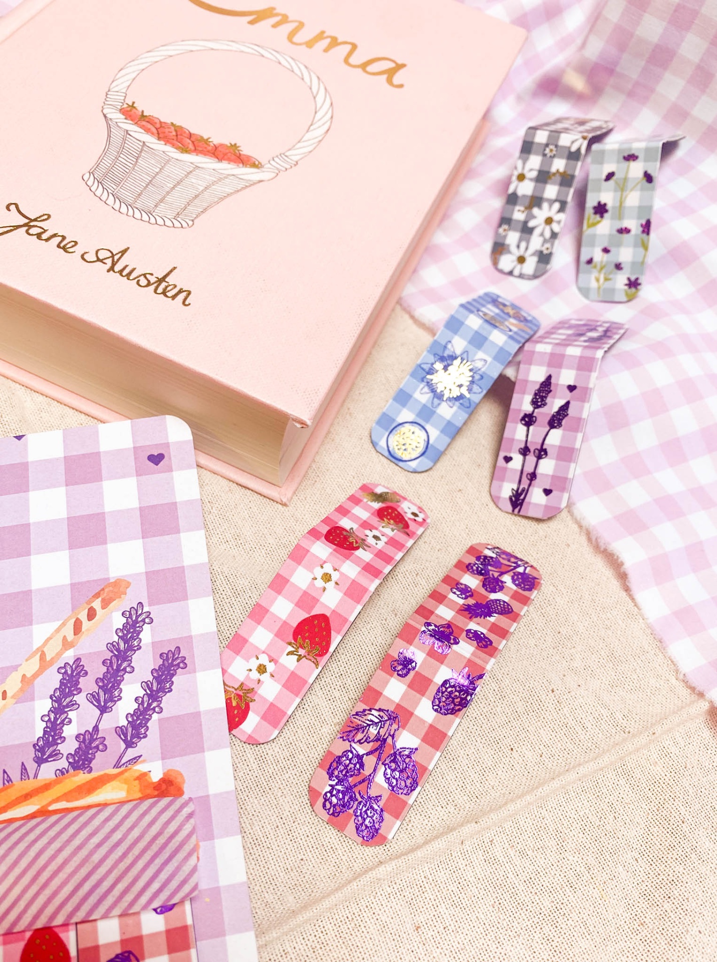 Marcadores de páginas metálicos coloridos com padrões xadrez e desenhos florais ao lado de um livro rosa intitulado EMMA por Jane Austen.