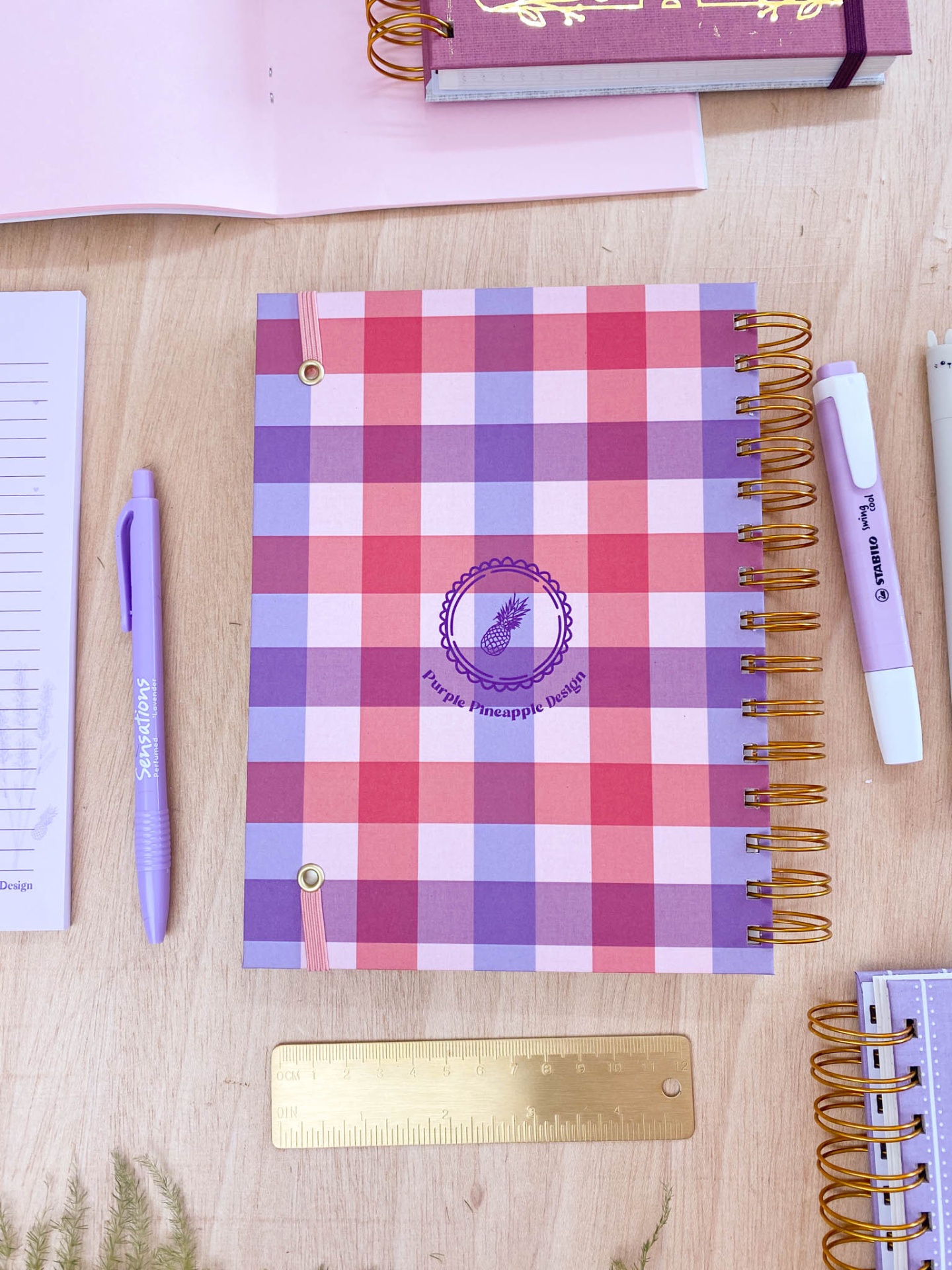 Caderno quadriculado vermelho rosa lilás branco com logótipo abacaxi e canetas sobre mesa de madeira