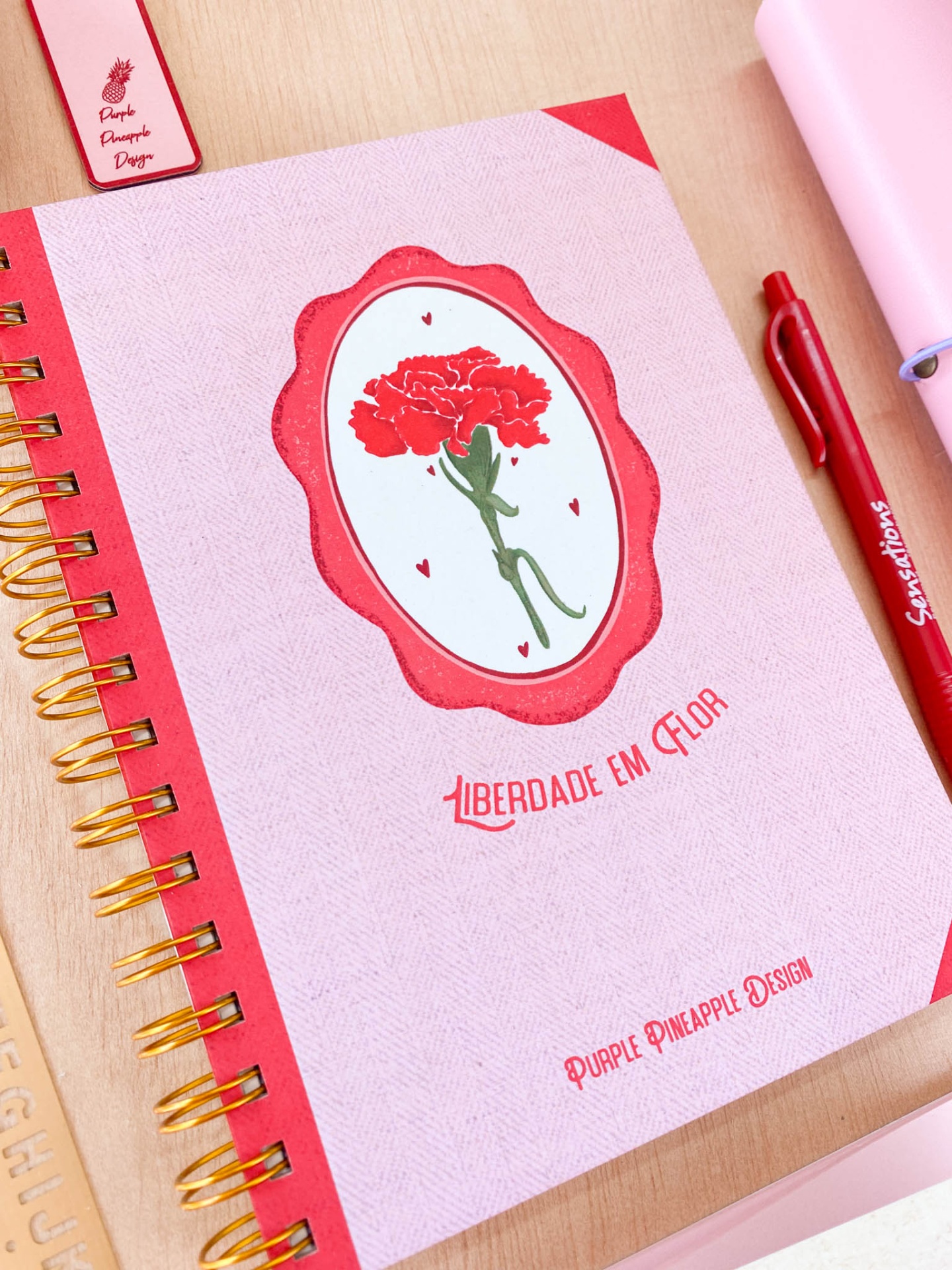 Caderno rosa com ilustração de flor vermelha e texto, caneta vermelha e artigos de papelaria sobre mesa de madeira.