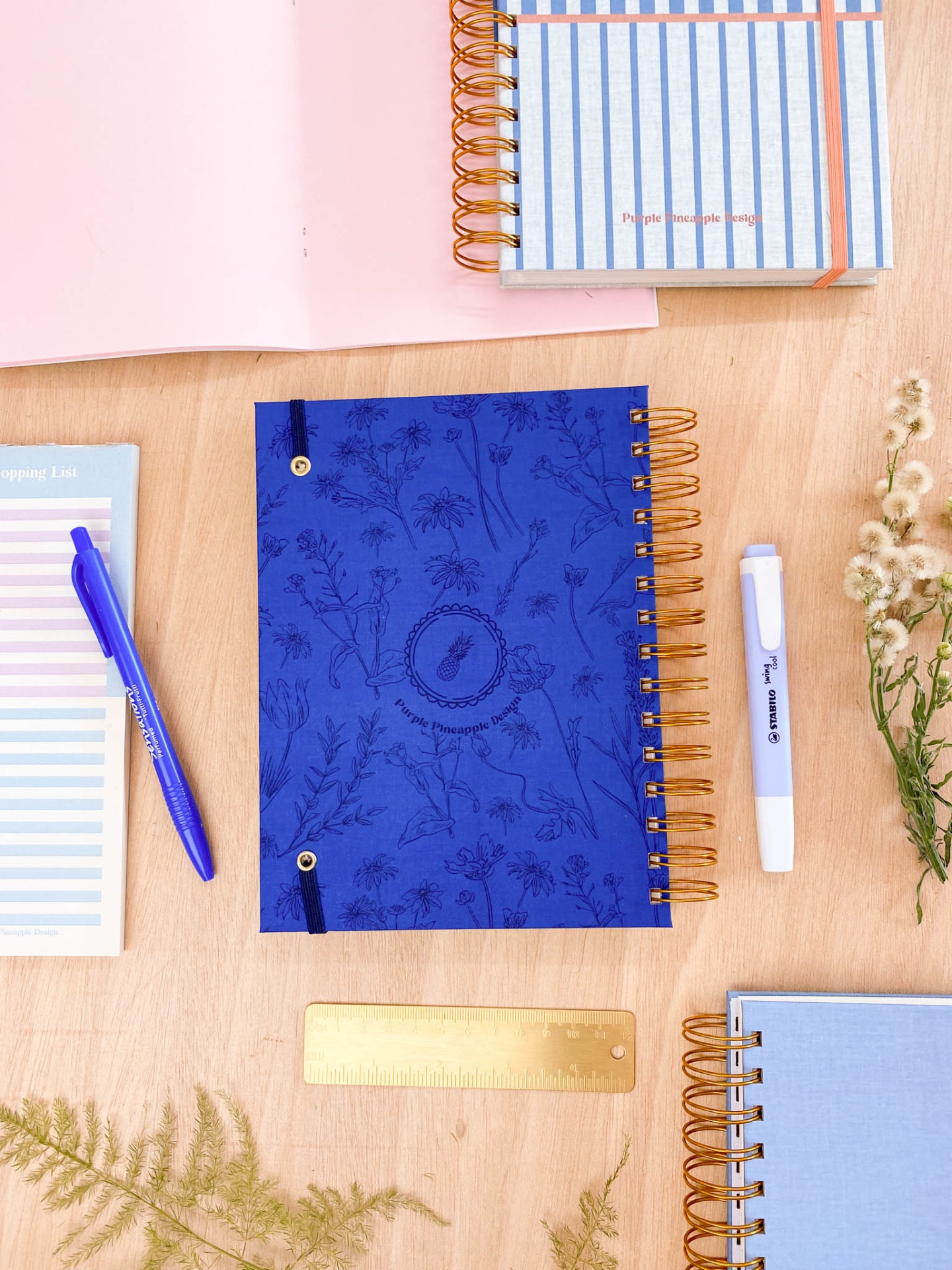 Caderno azul com padrão floral, caneta, marcador, régua e outro caderno sobre mesa de madeira