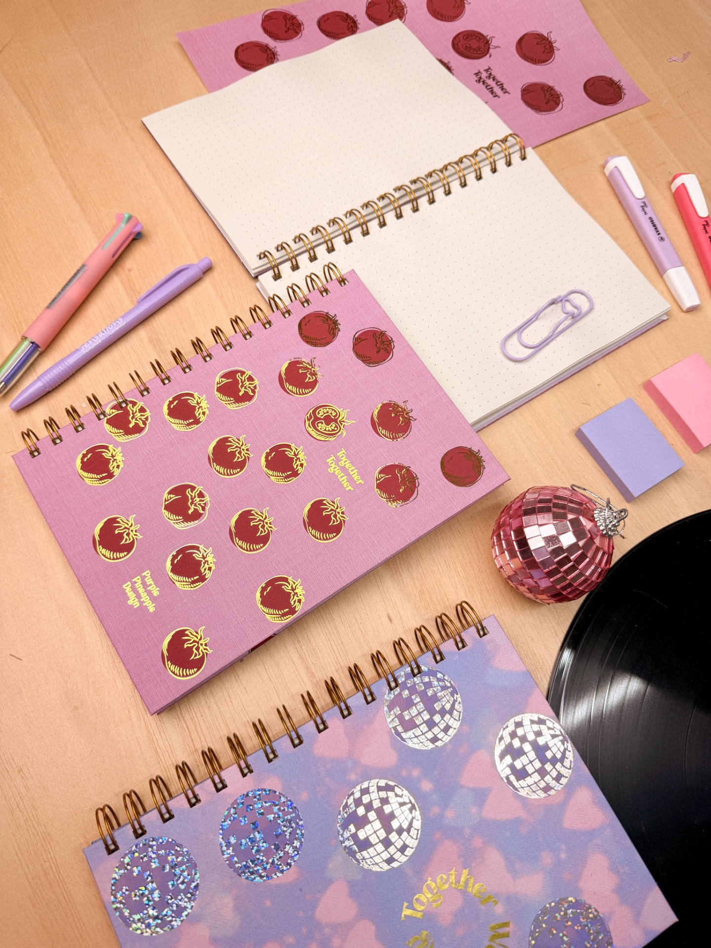 Cadernos espiralados decorativos com canetas, clips, post-its, bola de discoteca rosa e disco de vinil numa mesa de madeira