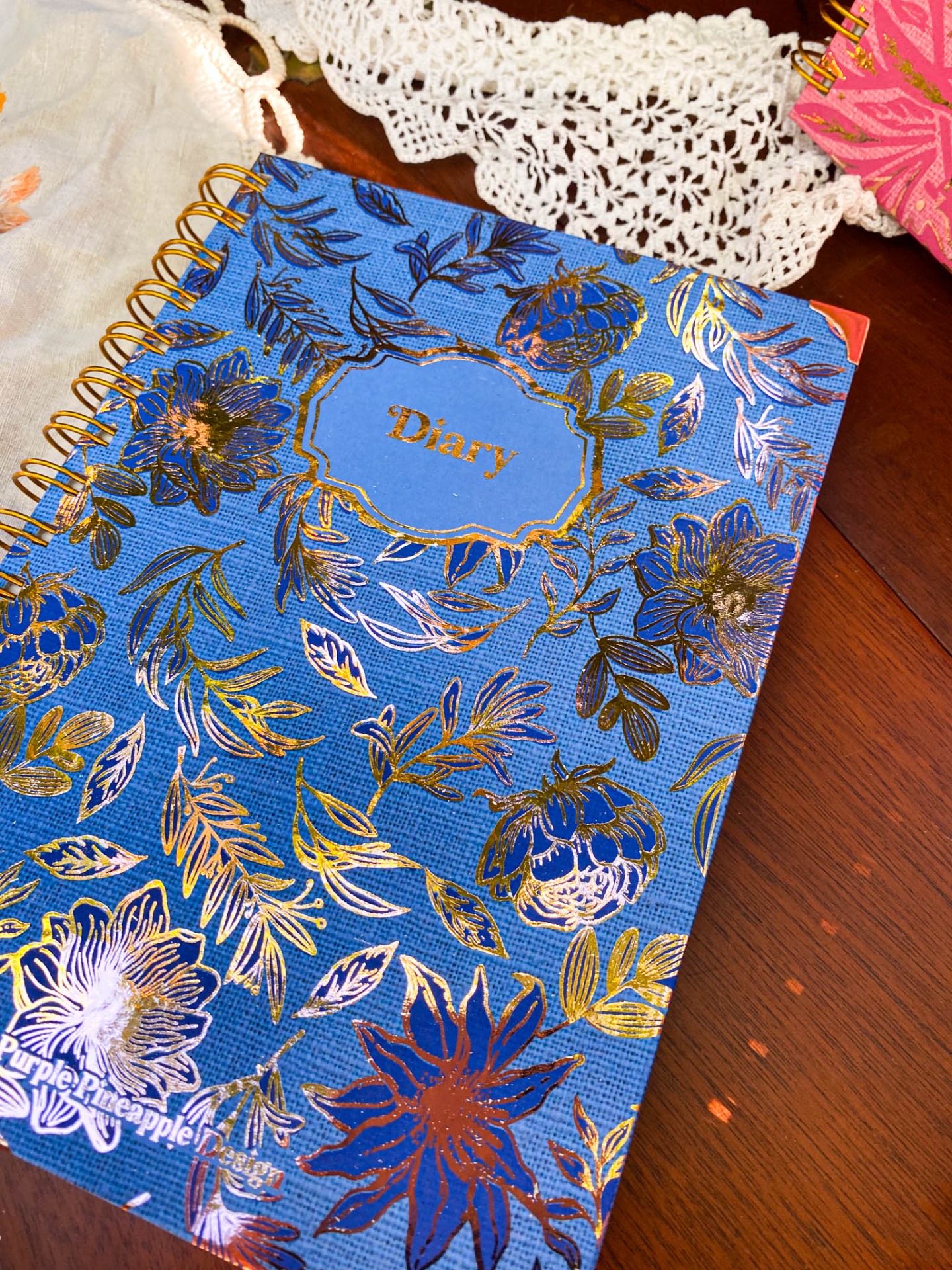 Caderno diário azul com flores douradas e espiral dourada sobre mesa de madeira junto a tecidos rendados