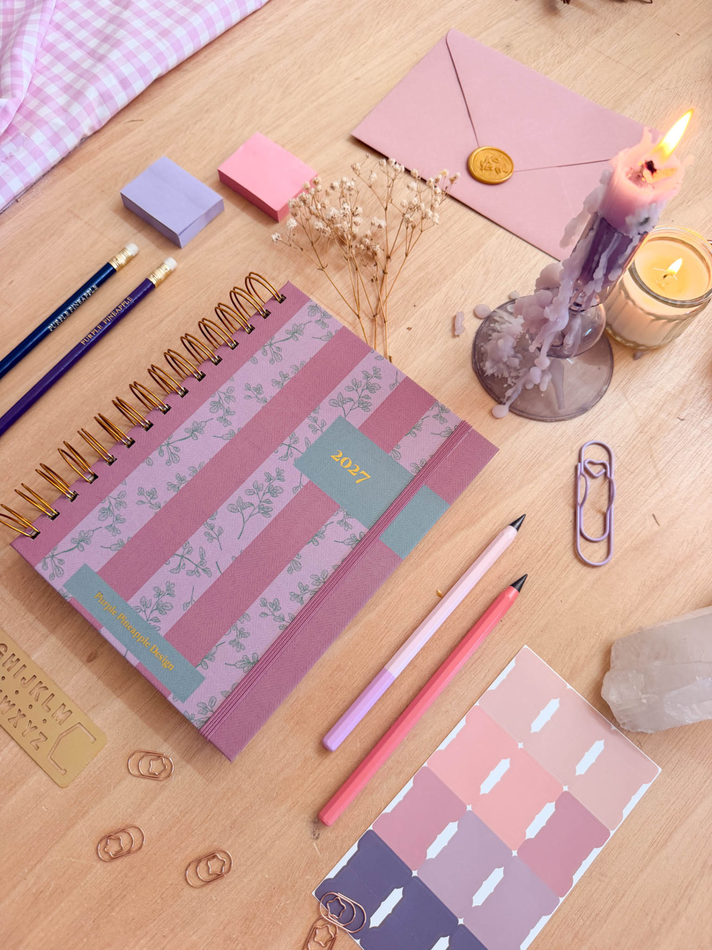 Mesa de madeira clara com agenda, lápis, envelopes, vela acesa e adesivos em tons rosa