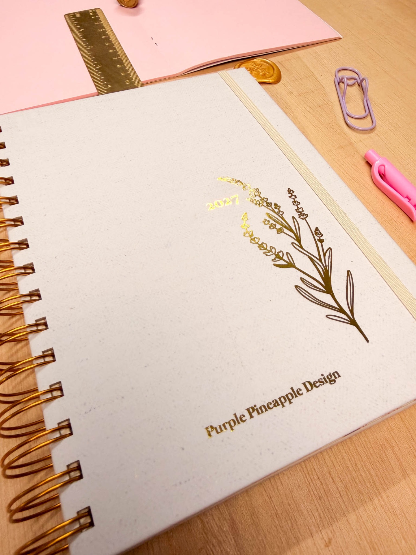 Caderno espiral branco com design dourado e texto, sobre mesa com régua, clips e caneta.