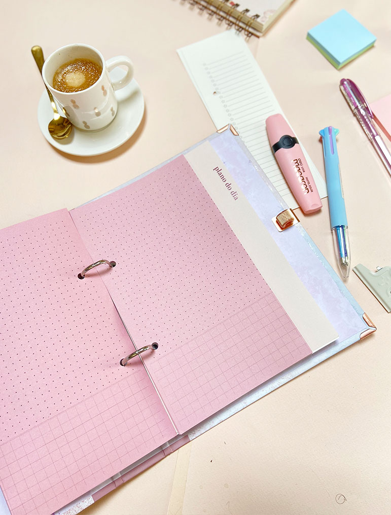 Agenda rosa aberta com canetas, marcador de texto, notas autocolantes e uma chávena de café sobre uma mesa clara.