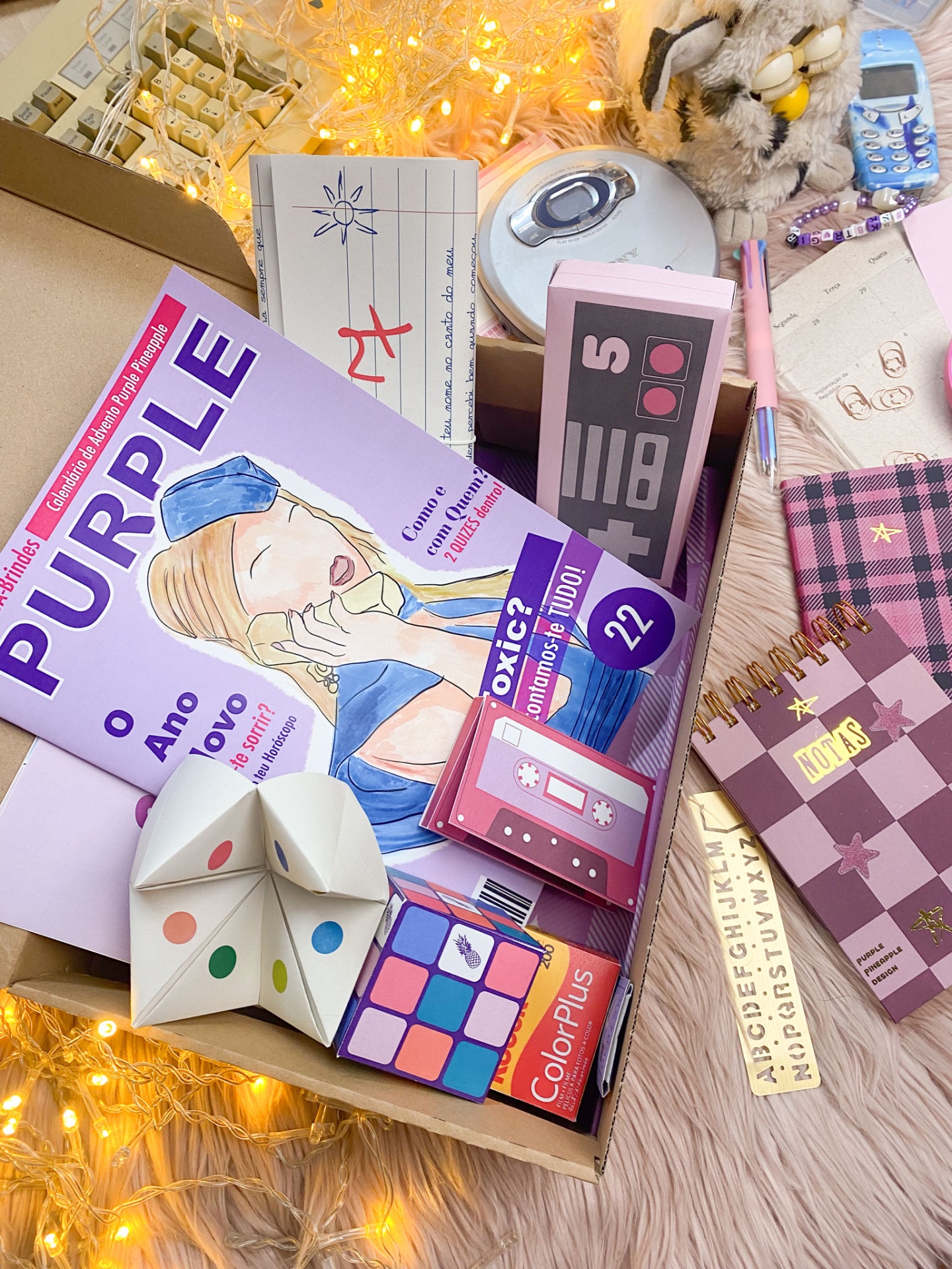 Caixa com artigos de papelaria como revista Purple, bloco de notas xadrez e brinquedo de papel colorido.