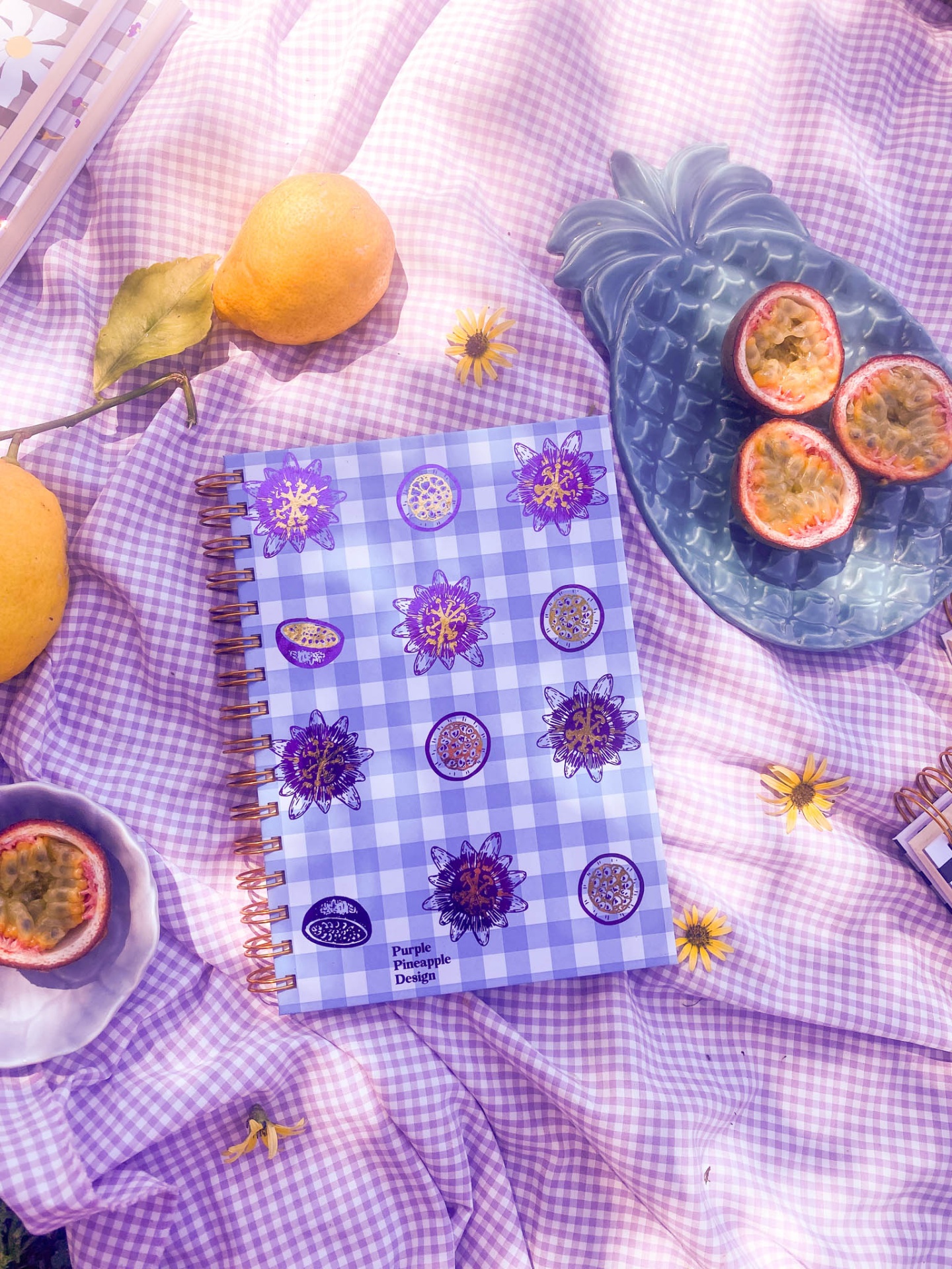 Caderno com capa xadrez roxa e branca e ilustrações, sobre tecido xadrez roxo com maracujás, limões e flores amarelas