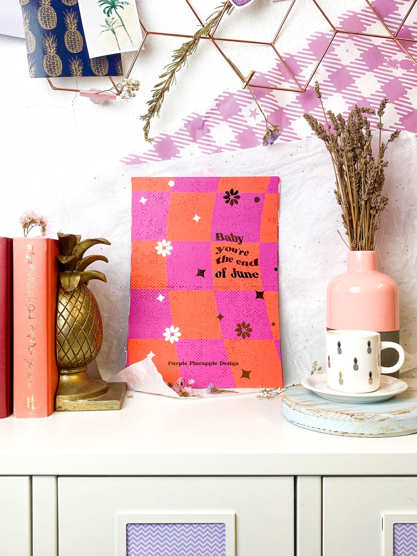 Conjunto decorativo com livro vermelho, estatueta dourada de ananás, cartaz rosa laranja, vaso rosa, chávena branca sobre superfície branca