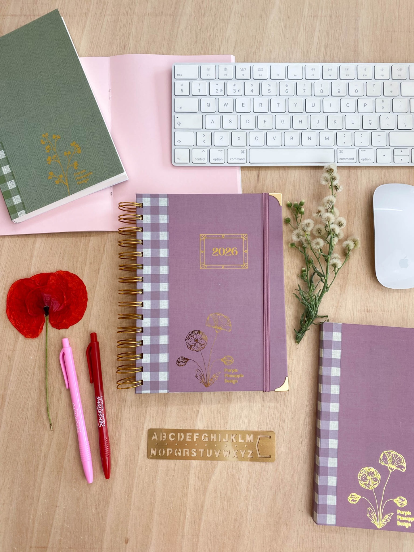Caderno roxo 2026 com padrão xadrez e detalhes dourados na mesa com teclado, canetas e flores