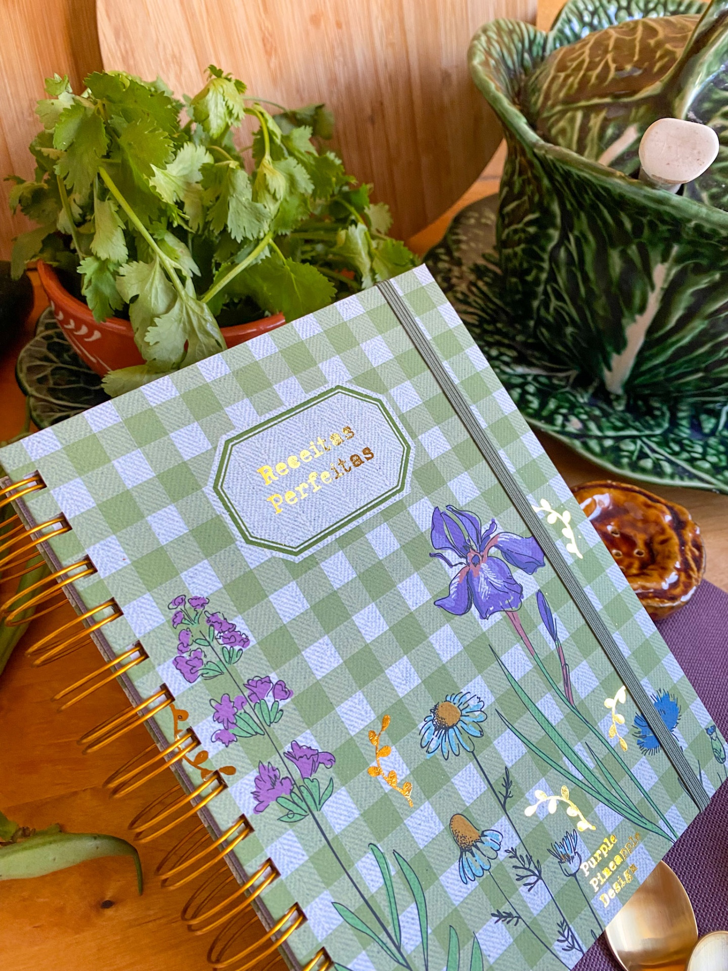 Caderno de receitas verde aos quadrados com flores e vaso de cerâmica verde com folhas.