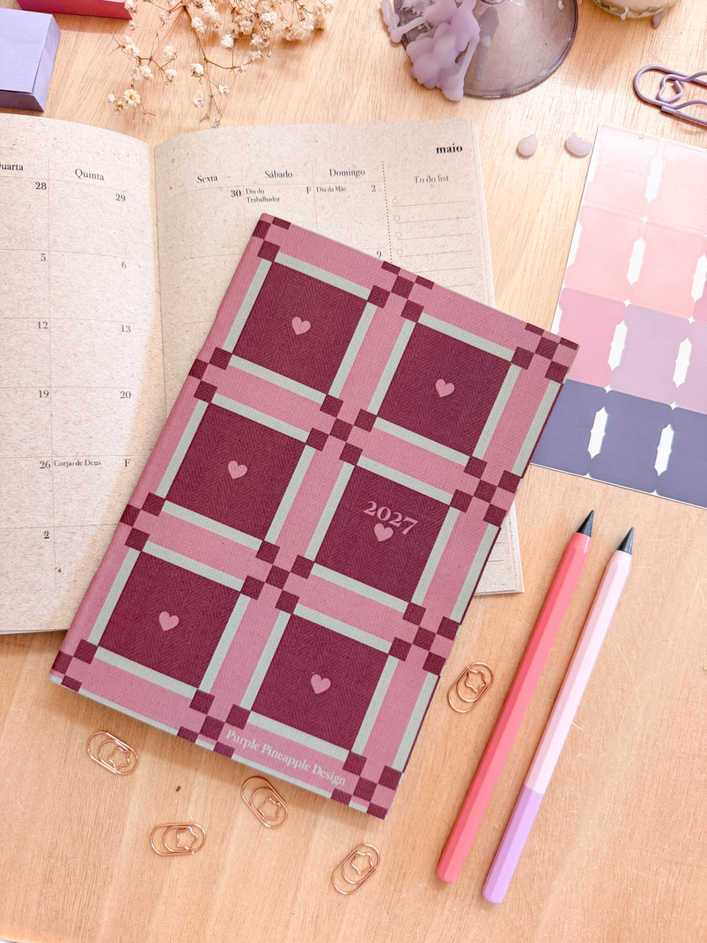 Agenda com padrão quadriculado rosa e vermelho, canetas e clipes em madeira clara