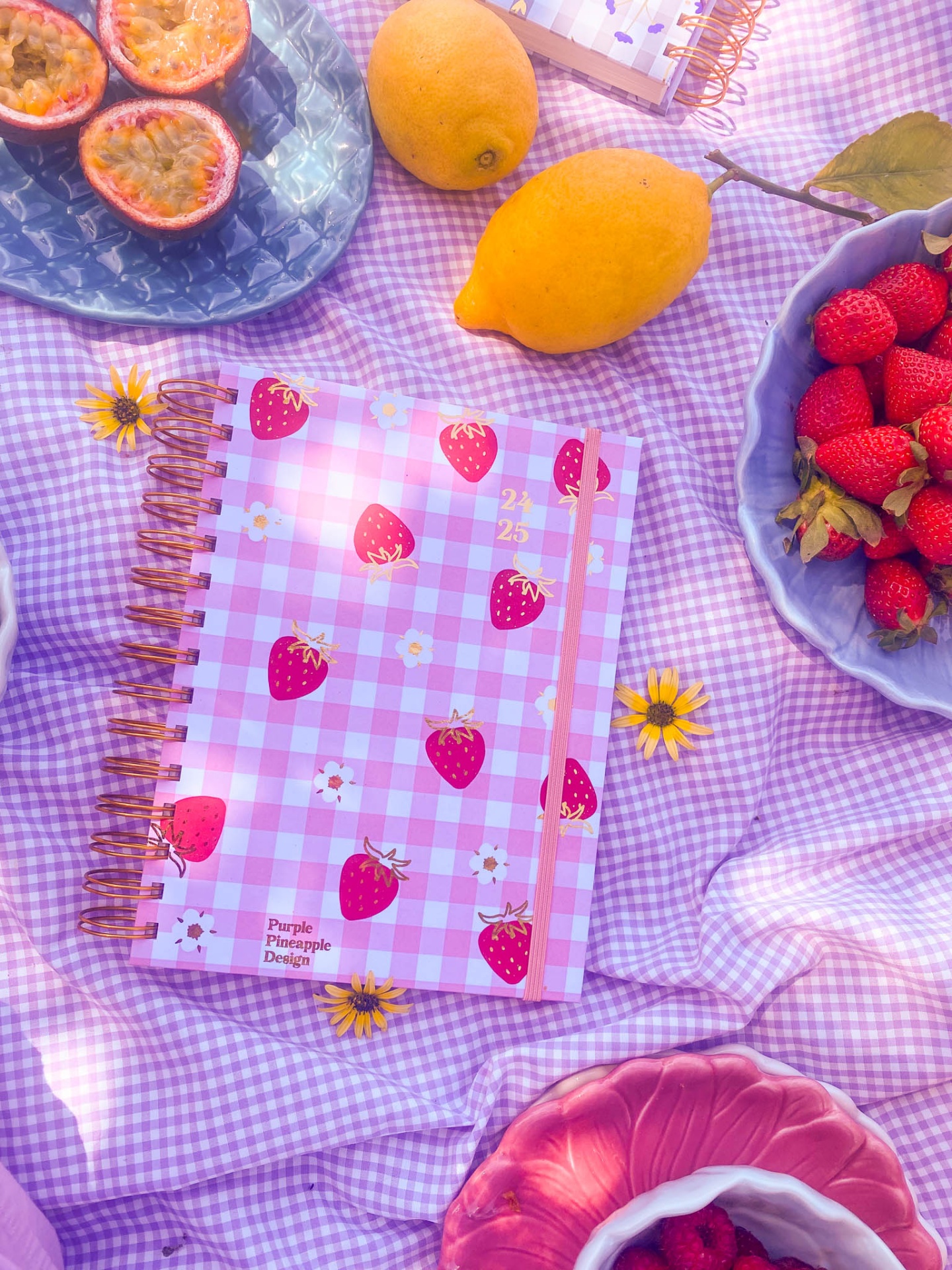 Caderno com capa rosa xadrez e ilustrações de morangos junto a frutas sobre tecido xadrez