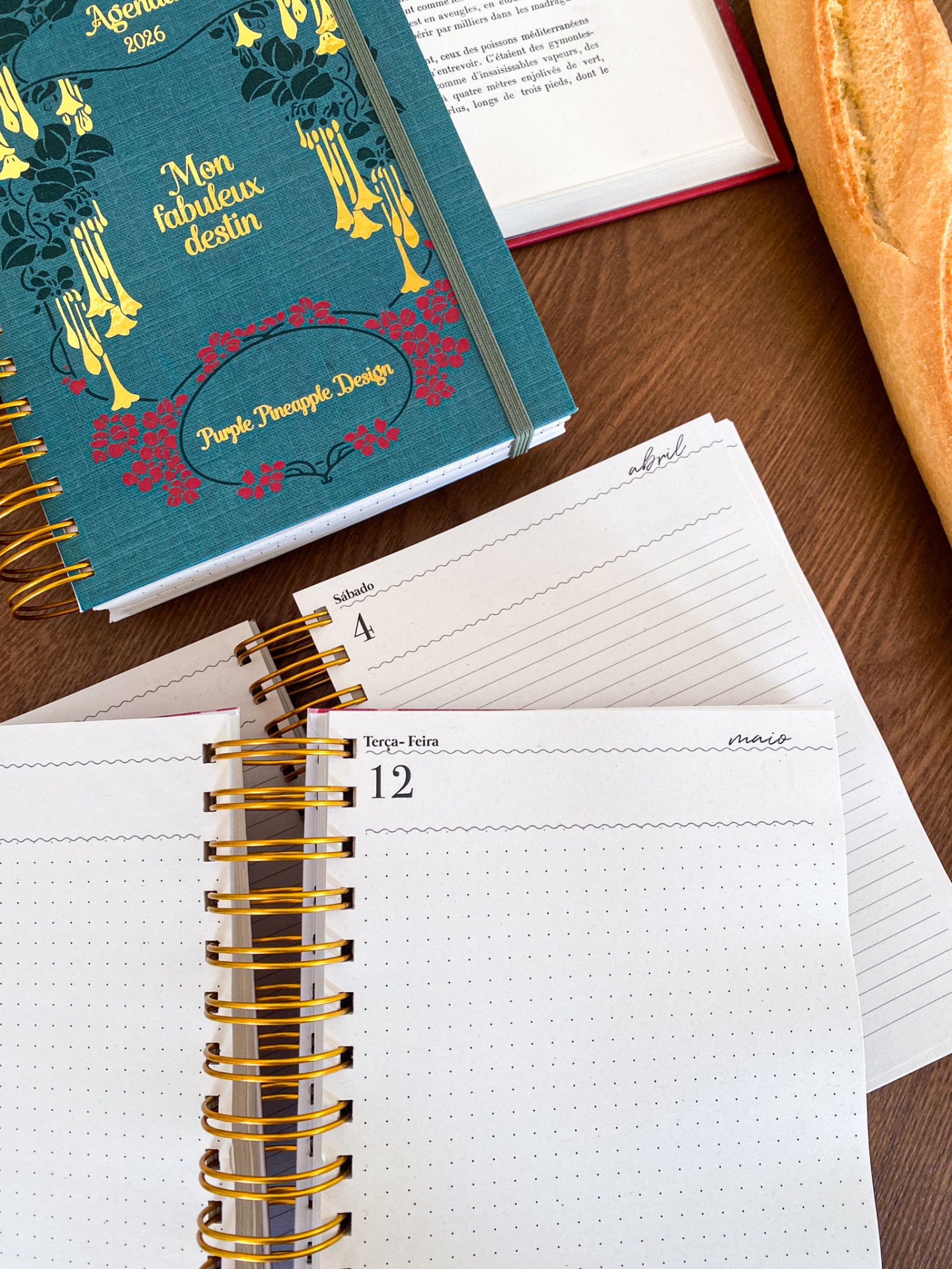 Agenda 2026 com capa azul decorada e páginas internas abertas numa mesa de madeira com baguete e livro aberto