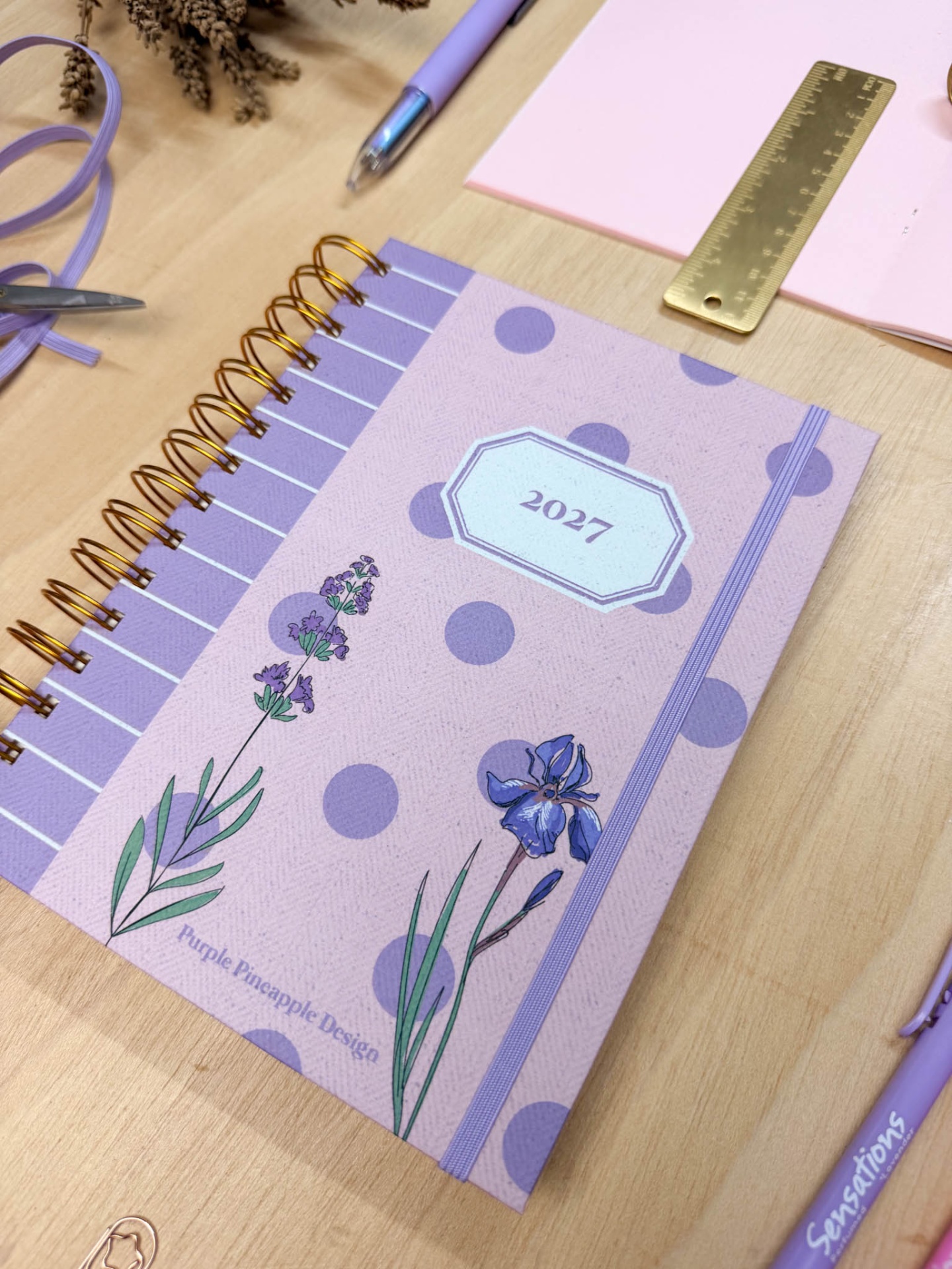Agenda lilás com flores e o ano 2027 numa mesa de madeira com caneta e régua