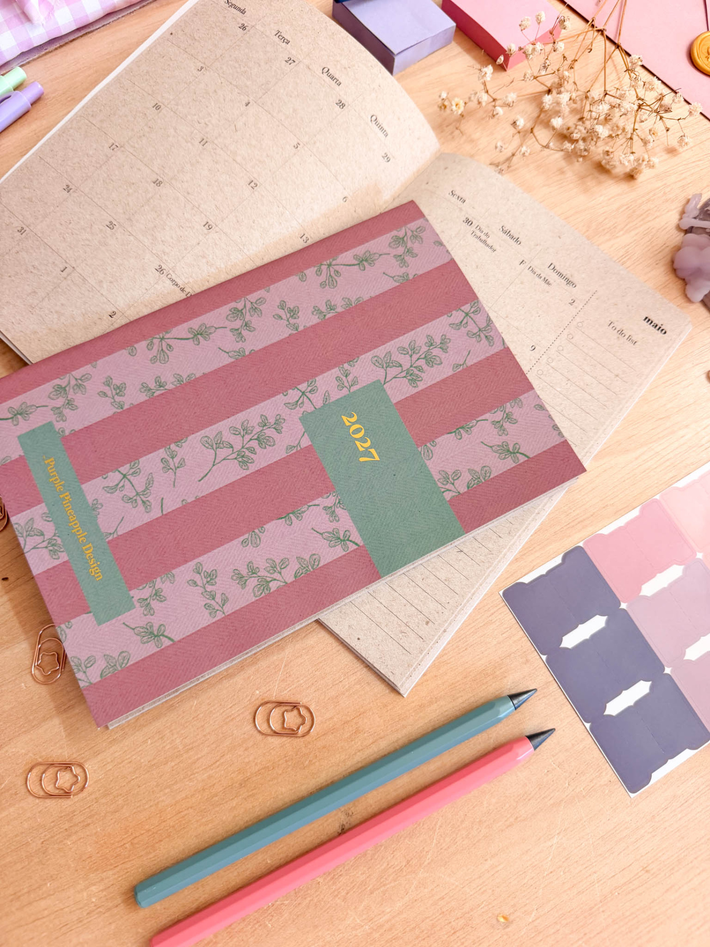 Agenda aberta de capa rosa com padrão floral em mesa de madeira com lápis, clips, autocolantes e flores secas