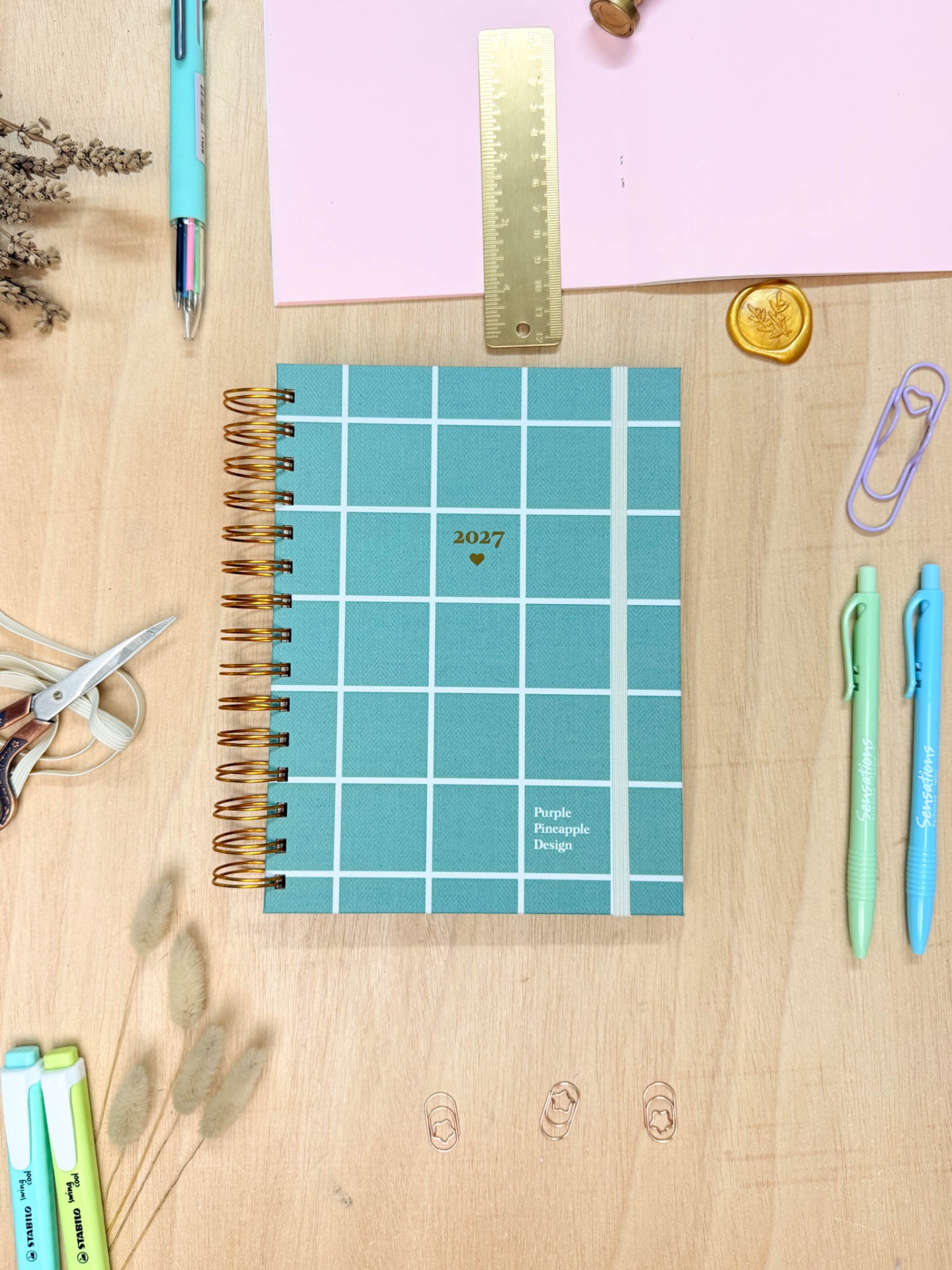 Agenda azul quadriculada com elástico branco e espiral dourada sobre mesa com papelaria colorida