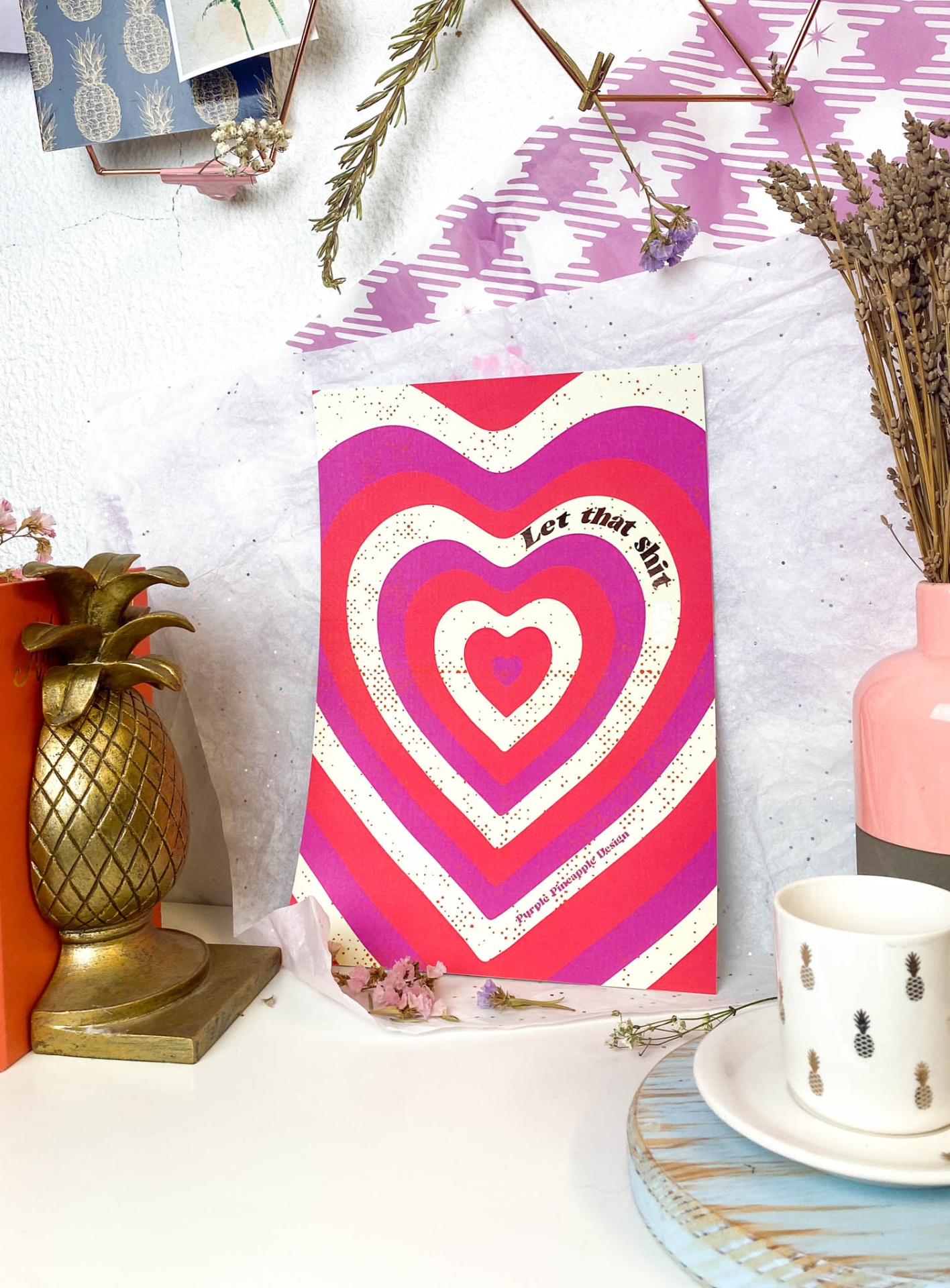 Livro com capa colorida em formato de corações, vaso rosa com flores secas, caneca branca com abacaxis, suporte dourado em forma de abacaxi numa bancada branca