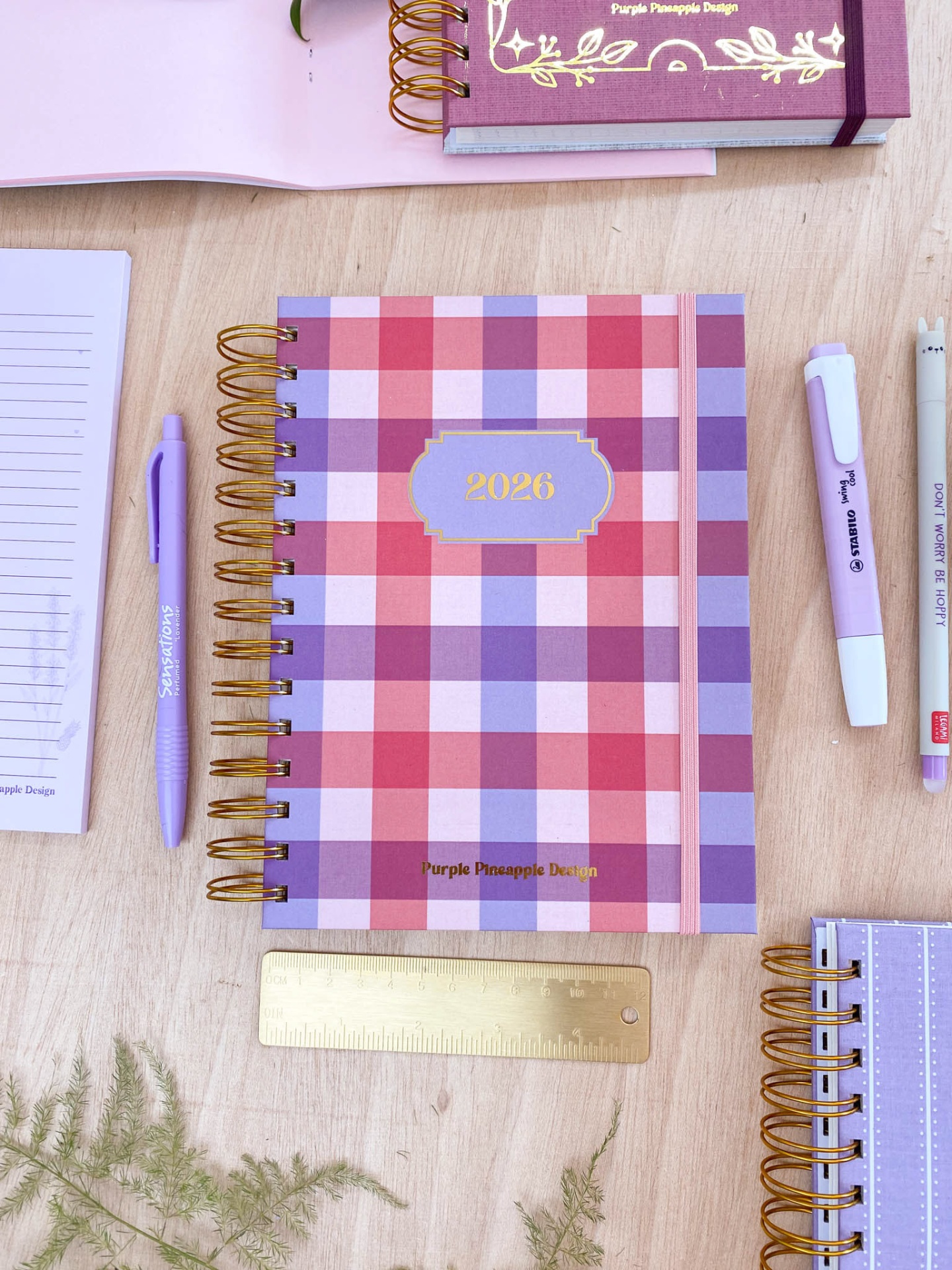 Caderno anual 2026 com padrão xadrez rosa e lilás e acessórios sobre mesa de madeira
