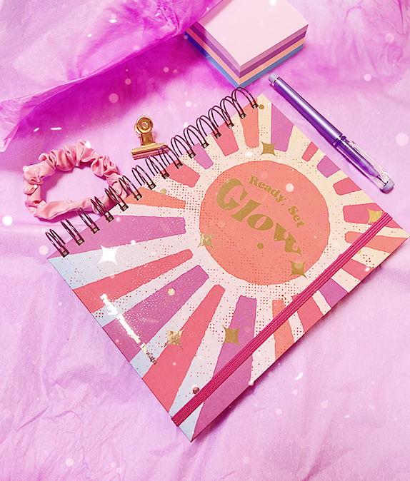 Caderno colorido com texto Ready Set Glow e acessórios de escritório em fundo rosa