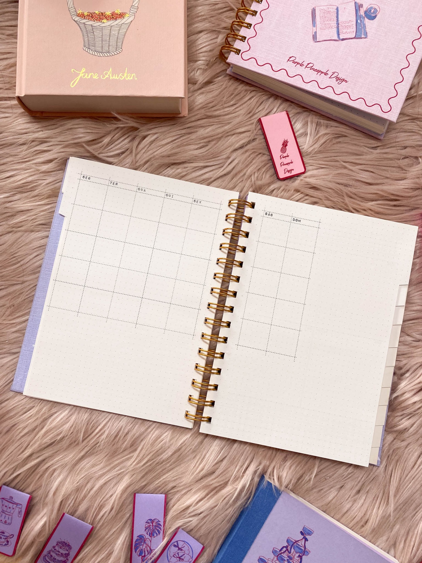 Caderno aberto com layout semanal e espiral dourado, acompanhado de outros artigos de papelaria em fundo rosa felpudo
