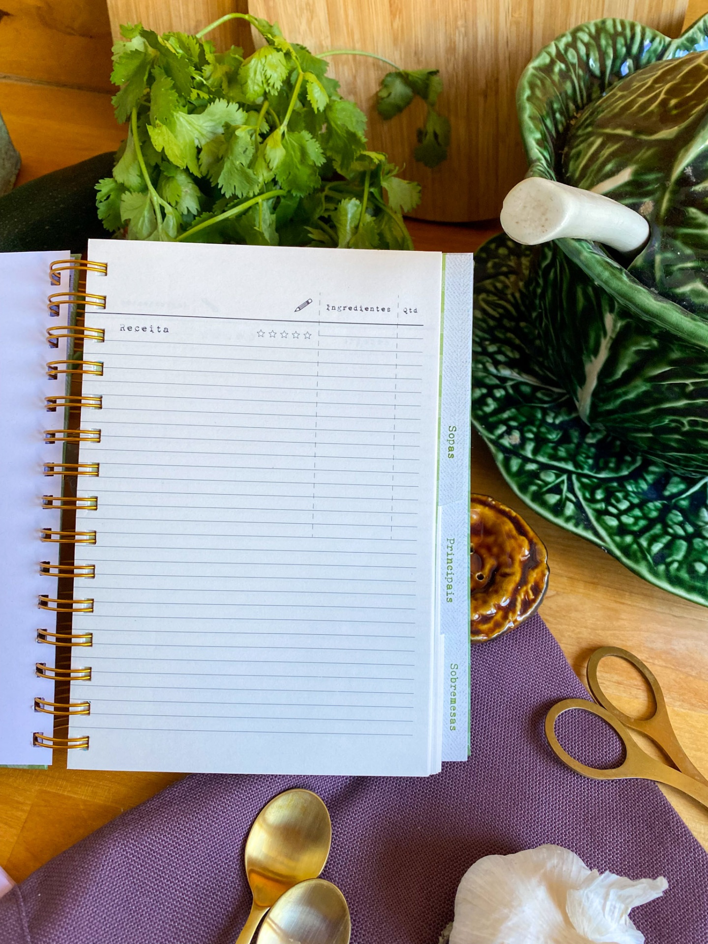Caderno de receitas aberto, coentros frescos, almofariz verde, colher dourada, alho e toalha roxa sobre mesa de madeira