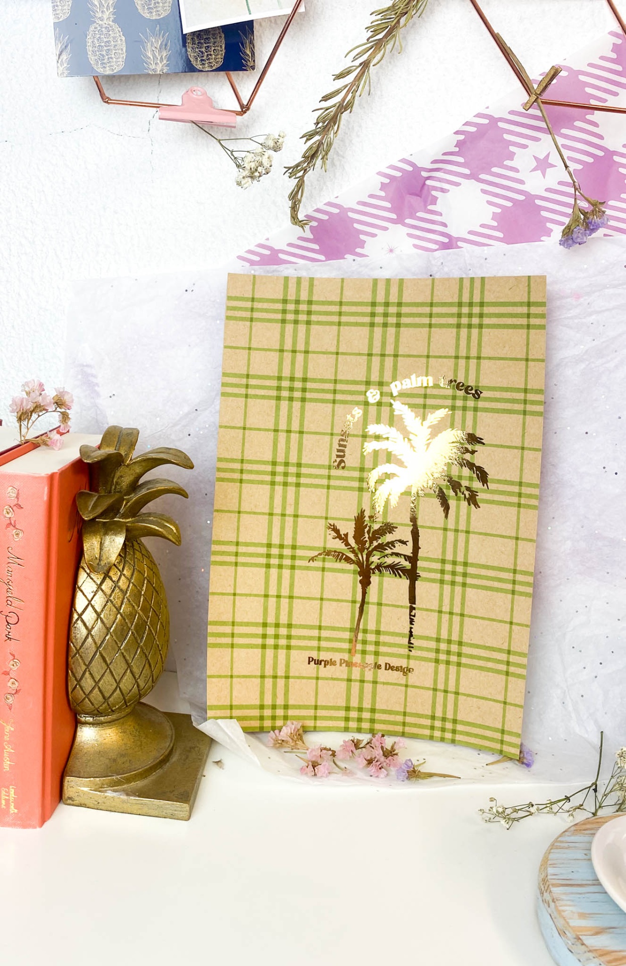 Caderno xadrez verde e bege com palmeiras douradas e texto, abacaxi decorativo e livro laranja numa mesa branca