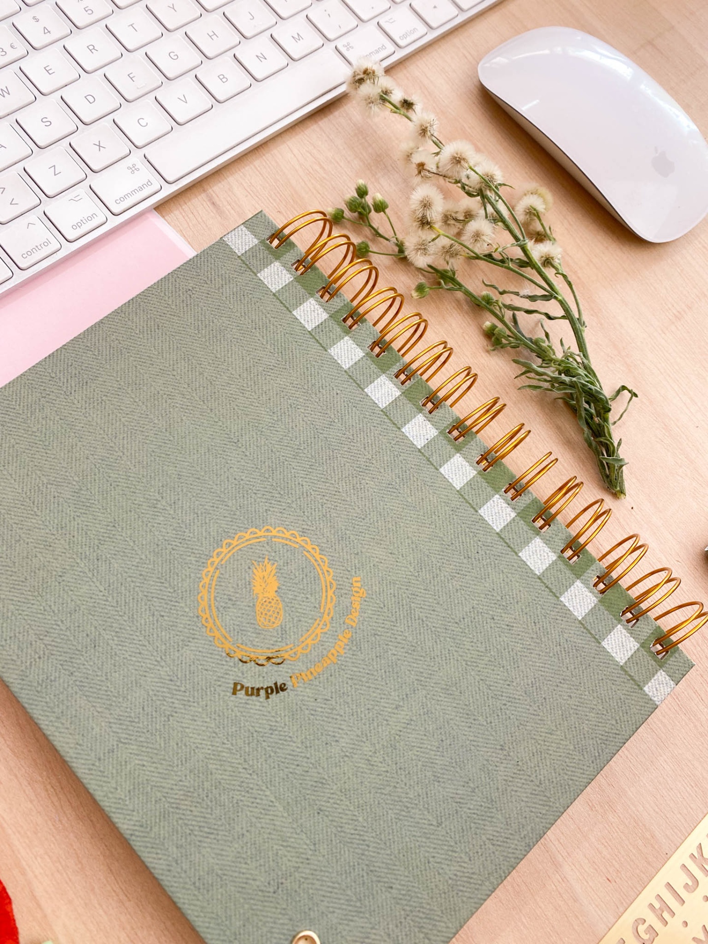 Caderno espiral verde com padrão xadrez e texto Purple, teclado white, e ramo de flores sobre mesa de madeira