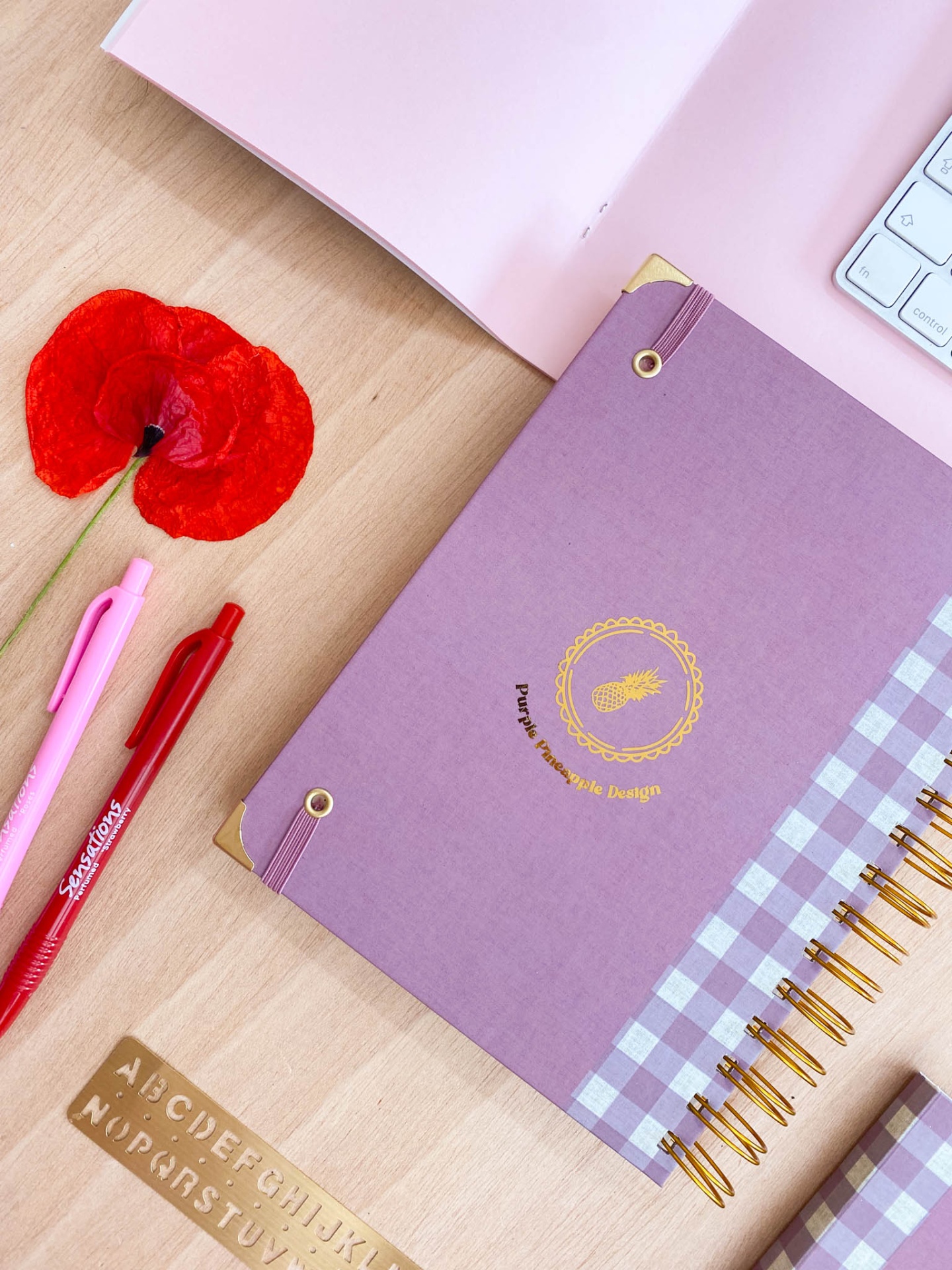 Agenda lilás com padrão xadrez, canetas, flor vermelha, teclado e régua de letras sobre mesa