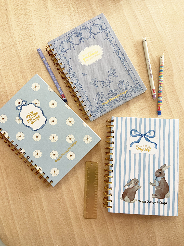 Três cadernos espiralados azuis com ilustrações e textos, lápis coloridos e régua dourada sobre mesa de madeira.