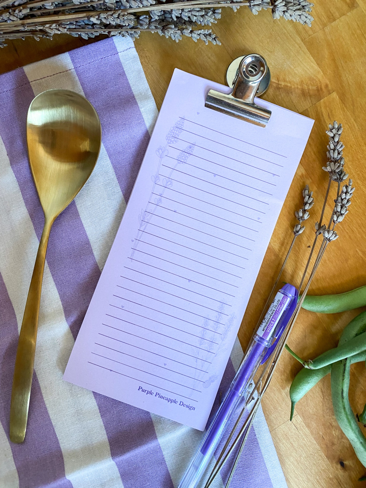 Bloco de notas com clip metálico sobre toalha riscada lilás e branco com colher dourada e caneta azul