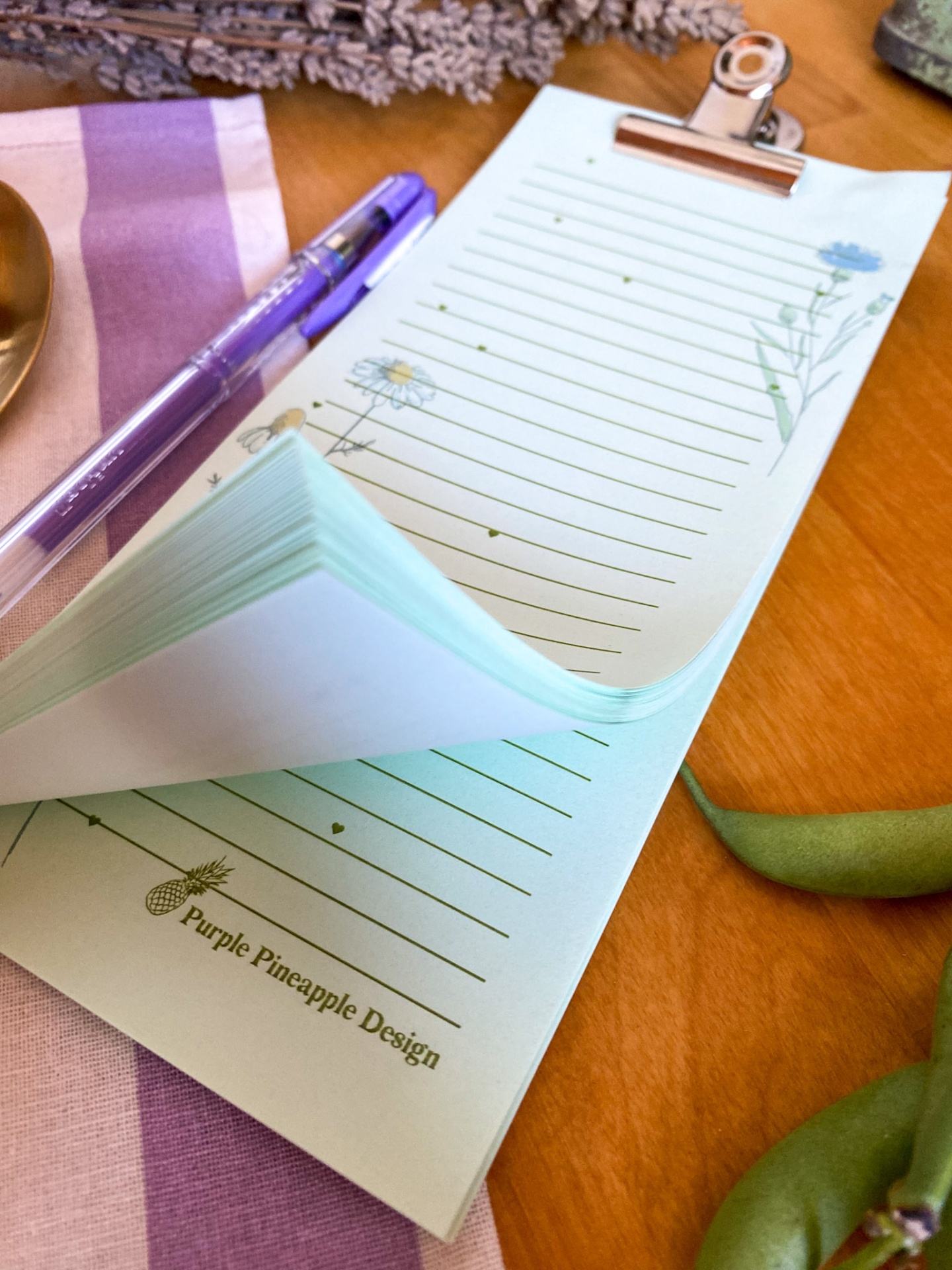 Bloco de notas com clip metálico e folhas decoradas com flores e linhas, caneta roxa translúcida, guardanapo lilás às riscas e planta verde sobre mesa de madeira.