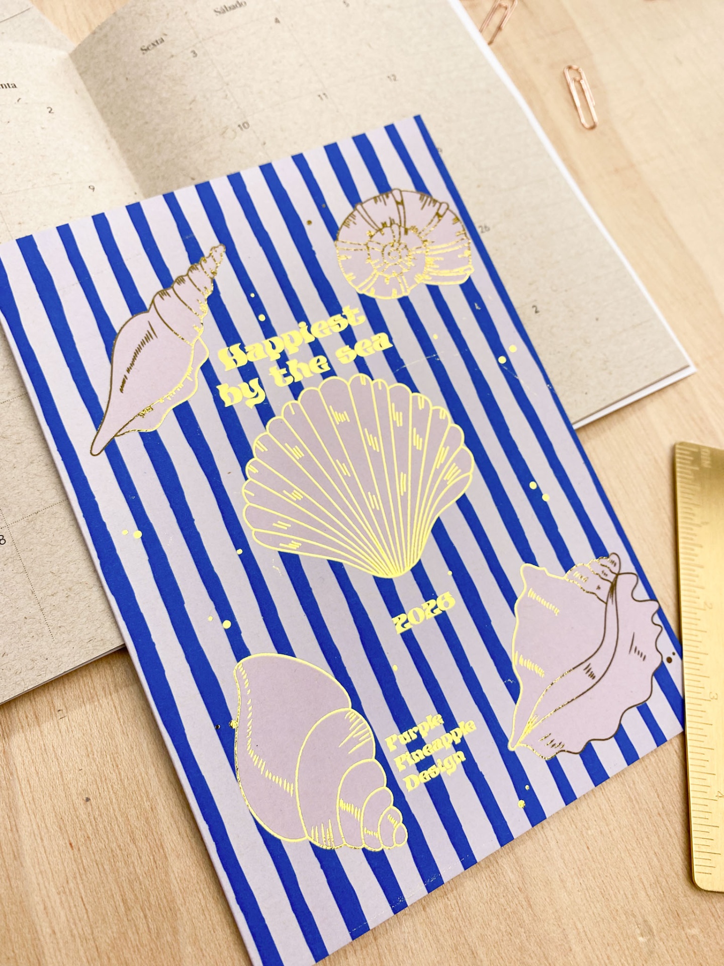 Caderno azul com riscas brancas e conchas douradas em mesa de madeira com calendário, clipe e régua