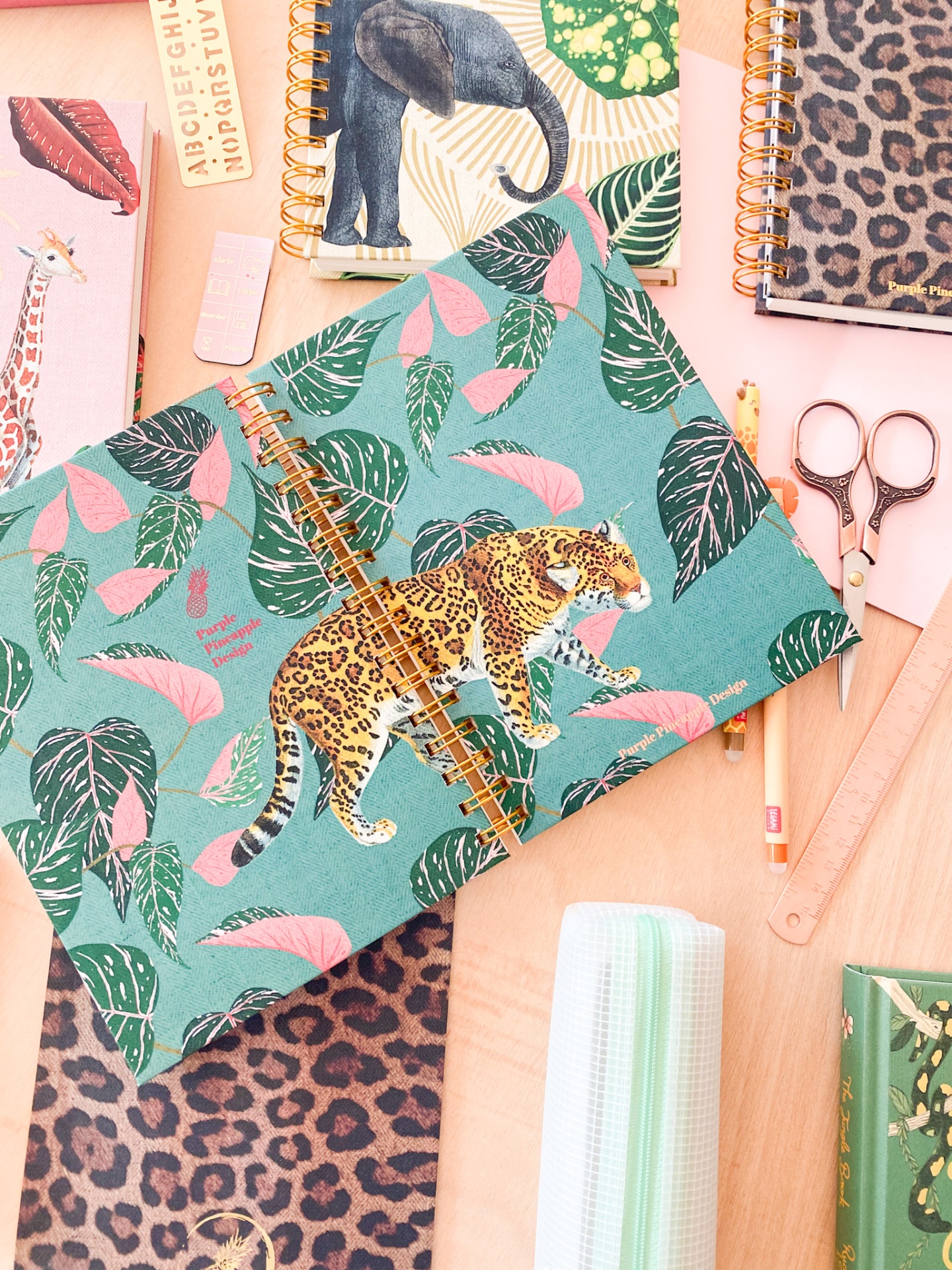 Cadernos espiral com capas coloridas de animais e folhas tropicais, acessórios de escritório em mesa de madeira.
