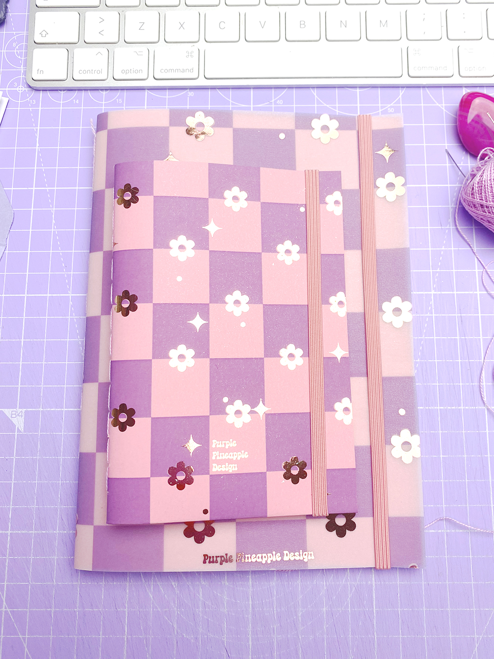 Cadernos rosa e roxo com padrão quadriculado e flores brancas, elástico rosa, sobre base lilás com teclado branco ao fundo