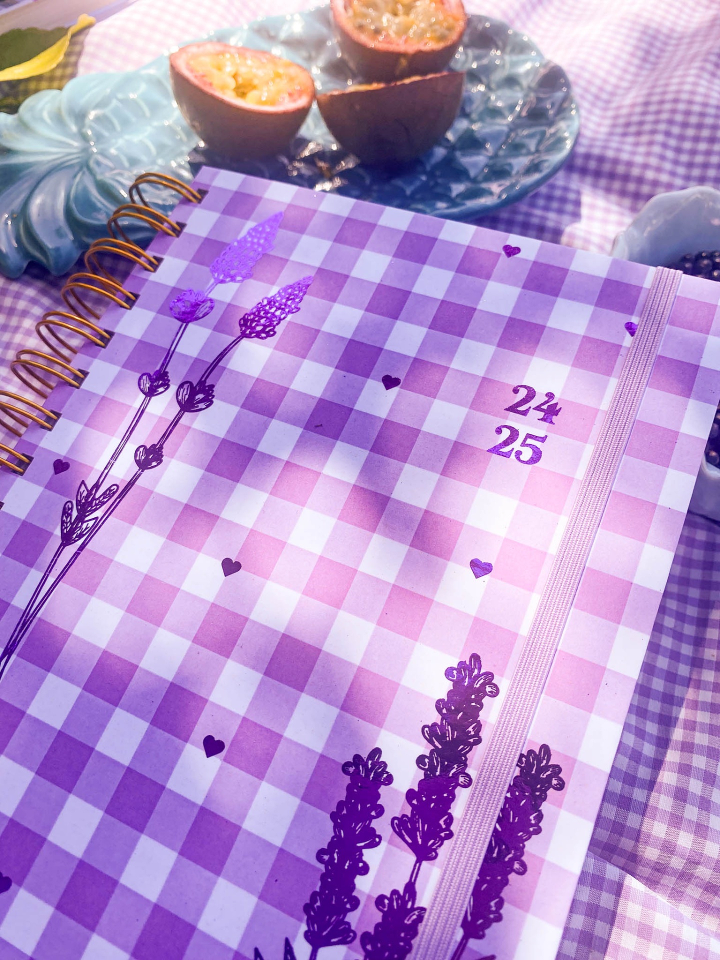 Caderno lilás xadrez com flores e números 24 e 25, ao lado de maracujás em prato azul