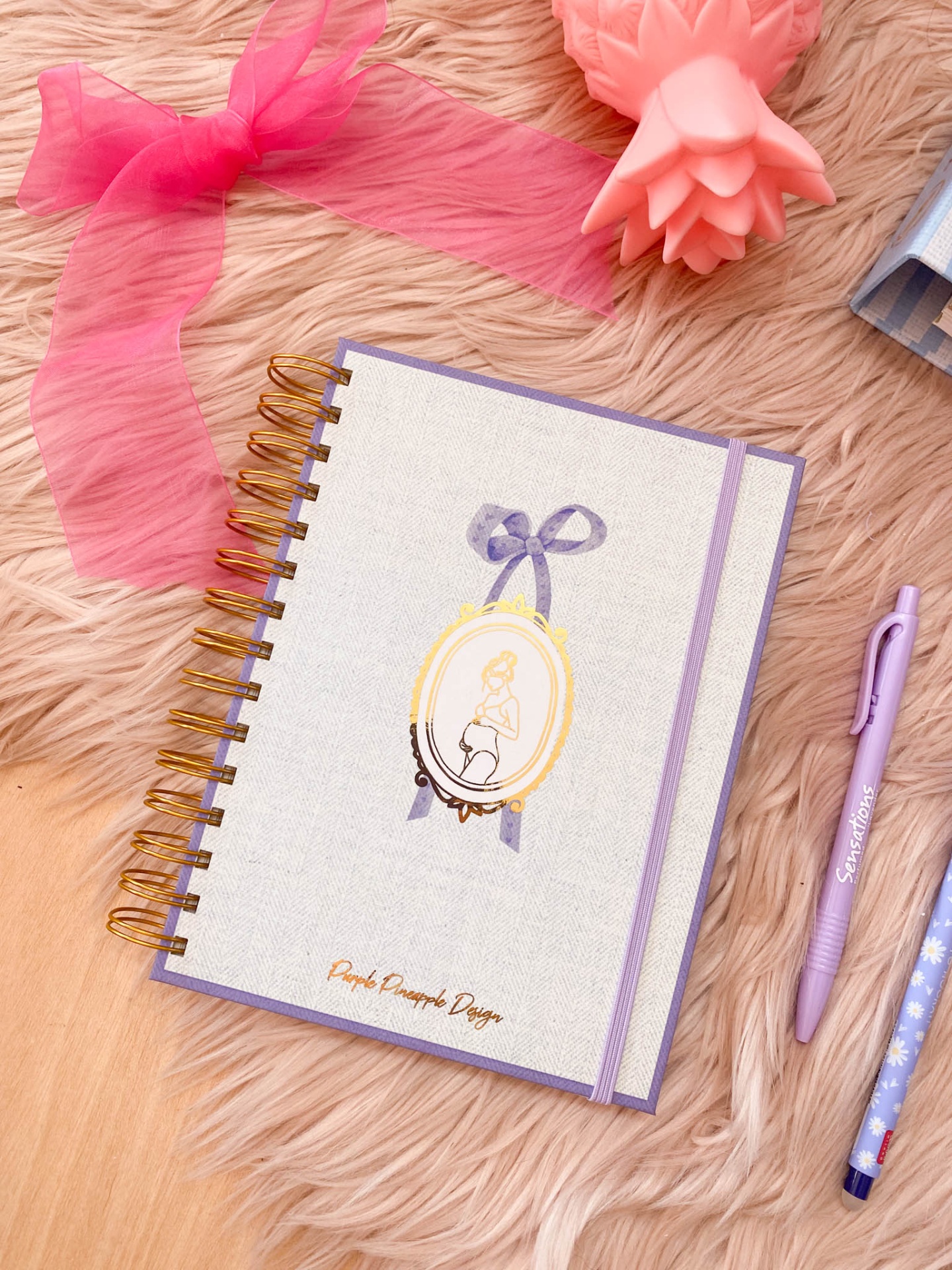 Caderno com capa branca e lilás com ilustração de mulher grávida e textos, sobre superfície em pelo rosa e madeira, rodeado por canetas, laço e vela.