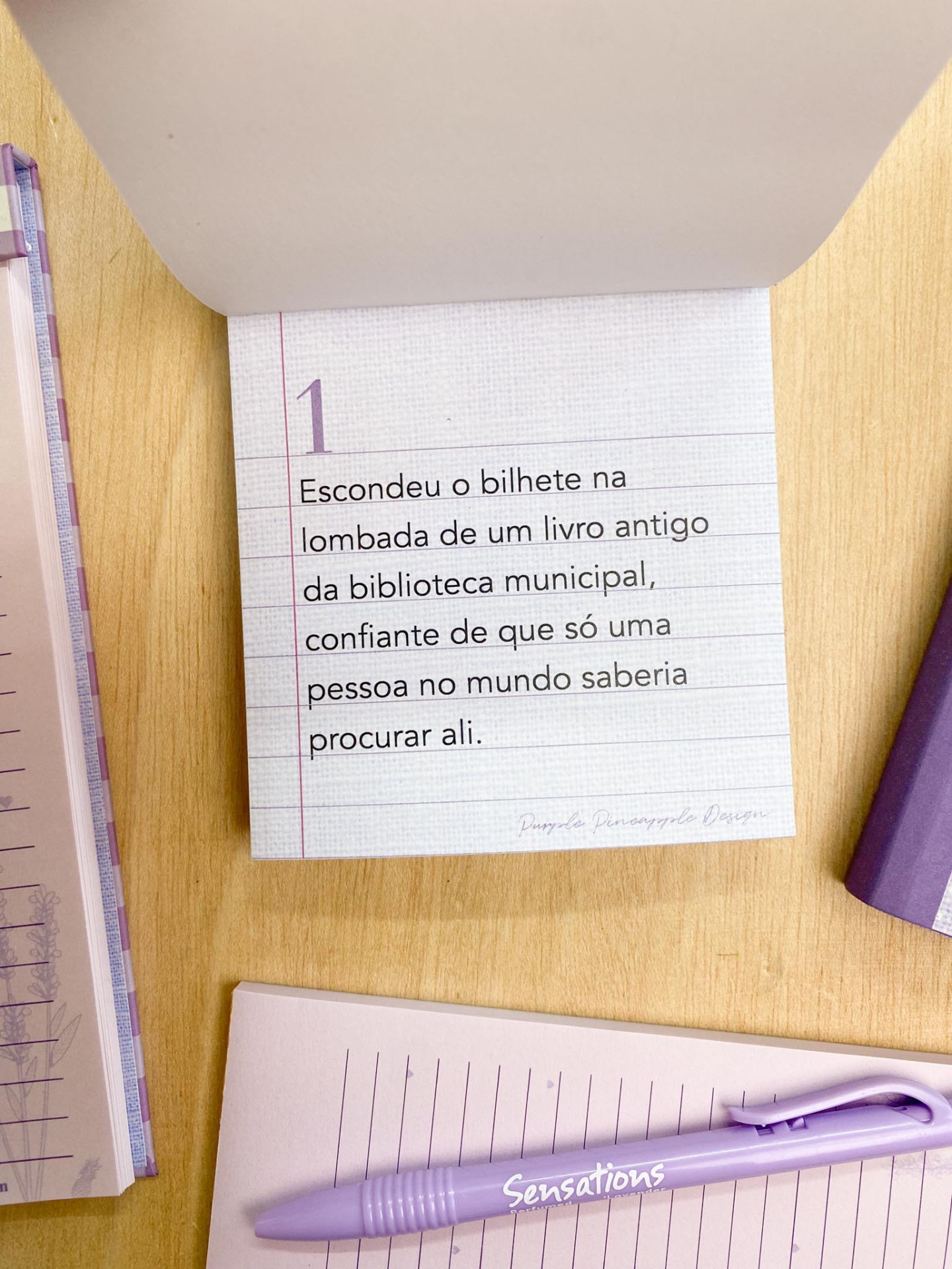 Bloco de notas com texto em português e caneta roxa numa mesa de madeira clara