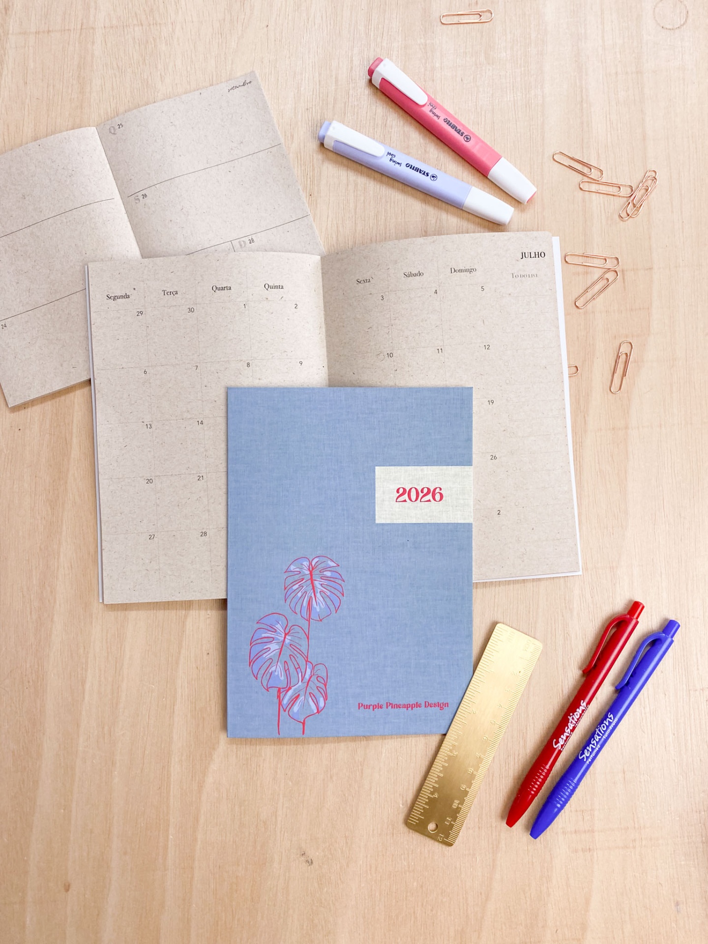 Agenda 2026 azul com ilustração vermelha, canetas, clips e marcador sobre mesa de madeira
