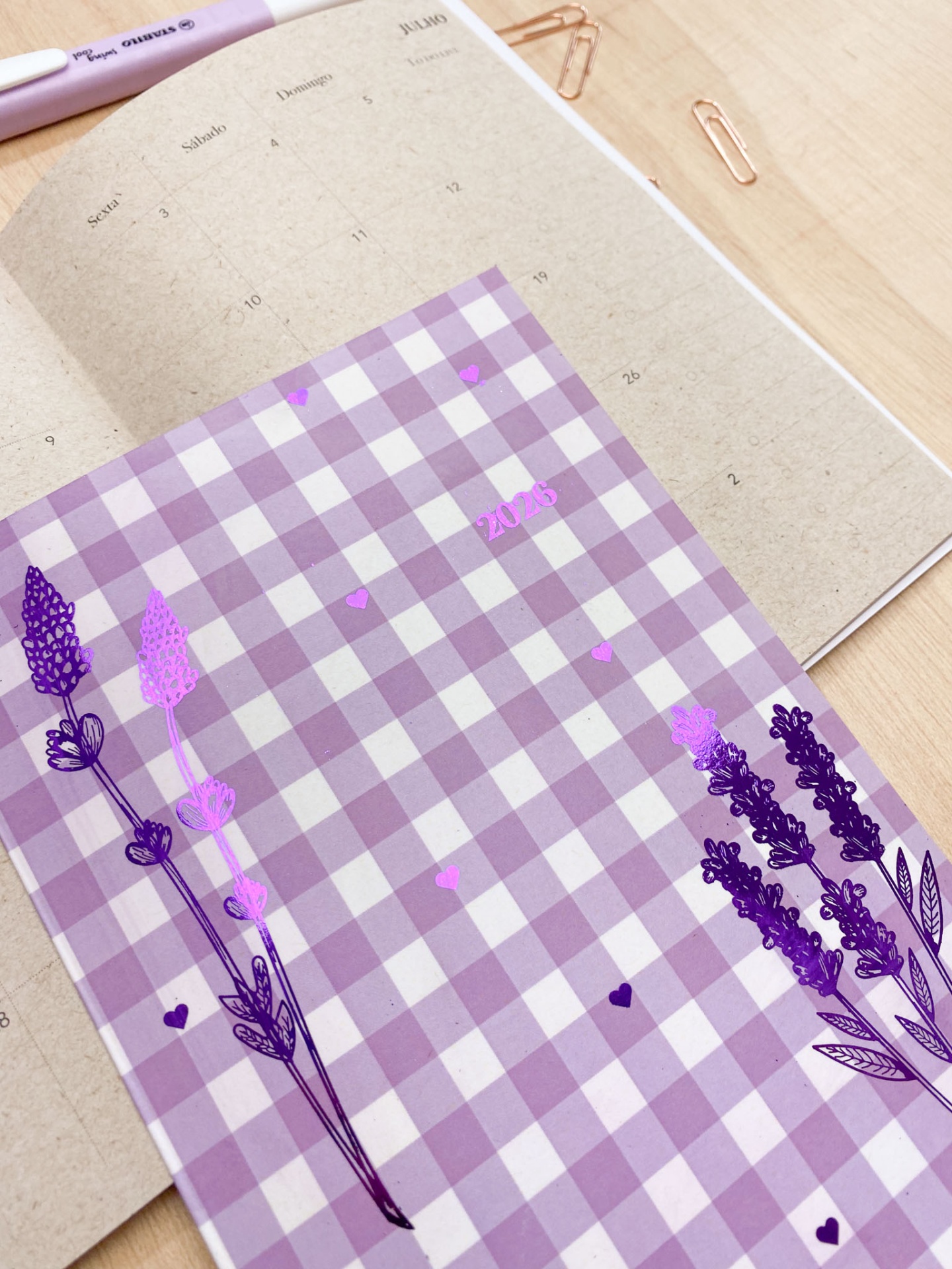 Calendário de mesa em papel kraft com capa lilás xadrez decorada com flores e a palavra 2023, caneta rosa e clips dourados sobre mesa de madeira clara