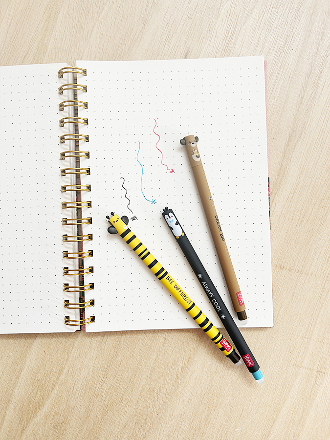 Três canetas decorativas coloridas sobre caderno espiral aberto com folhas pontilhadas.