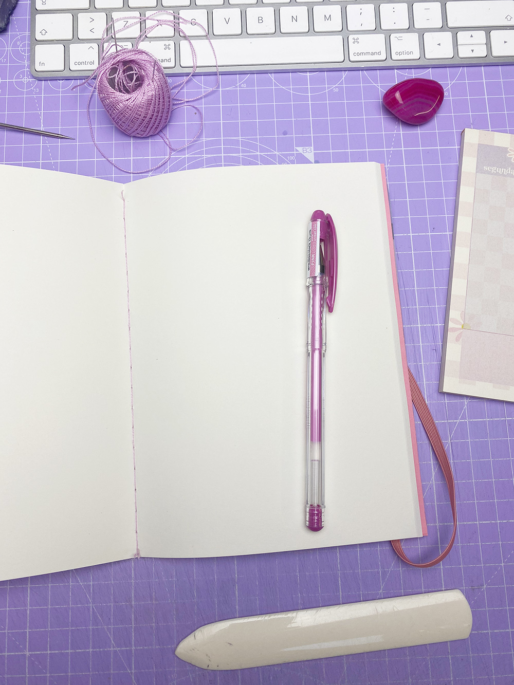 Caderno rosa aberto com caneta roxa sobre uma mesa lilás quadriculada e vários itens de escrita.