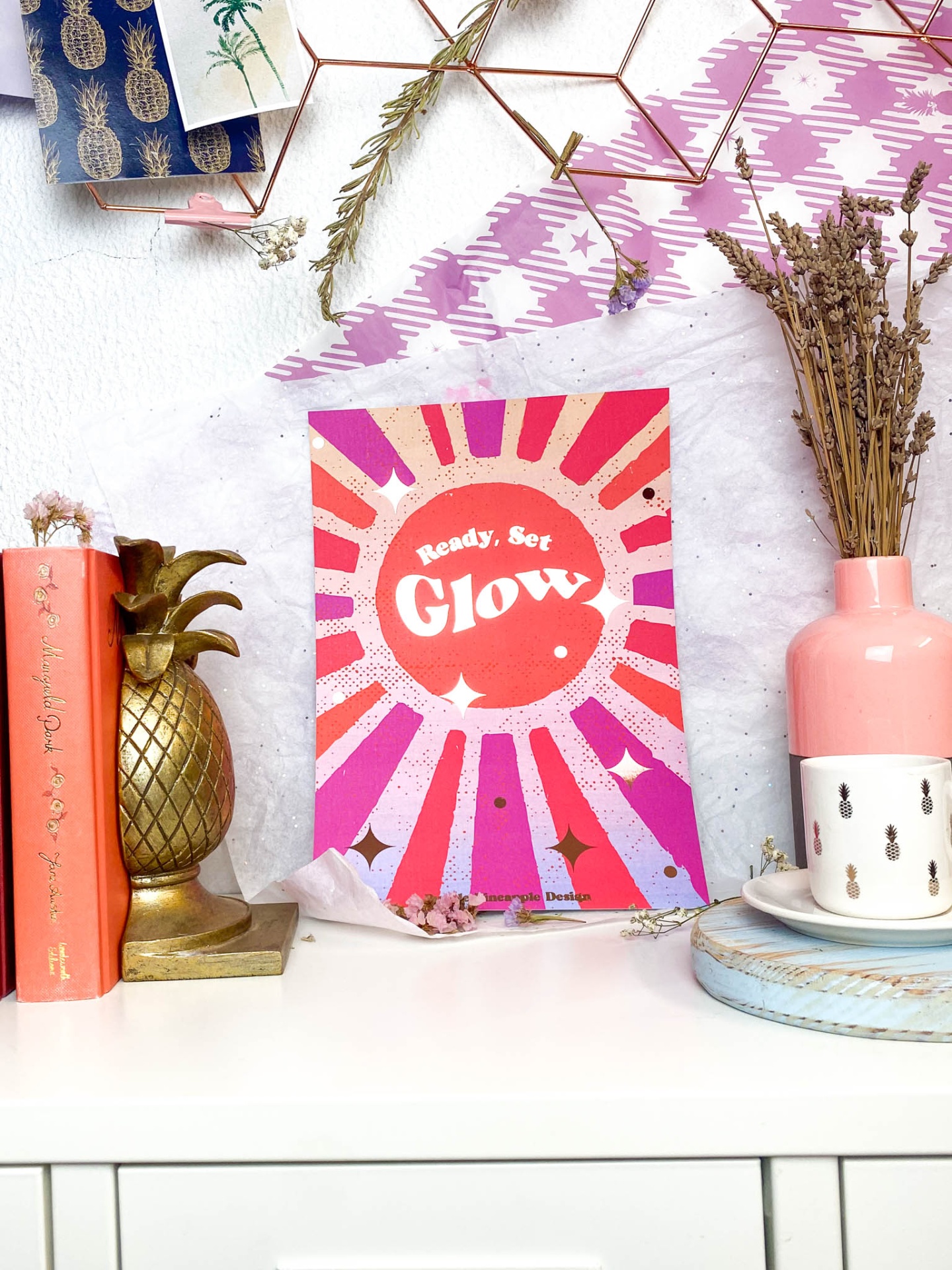 Cartaz colorido com as palavras 'Ready, Set Glow', livro pêssego, suporte abacaxi dourado e vaso rosa com flores secas