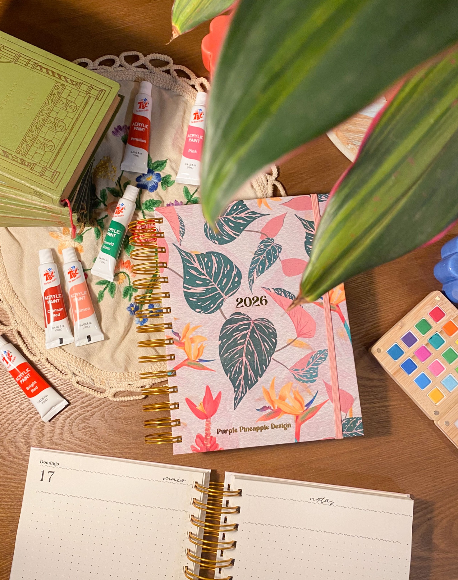 Agenda 2026 floral, tintas acrílicas, livro verde e paleta colorida sobre mesa de madeira com plantas