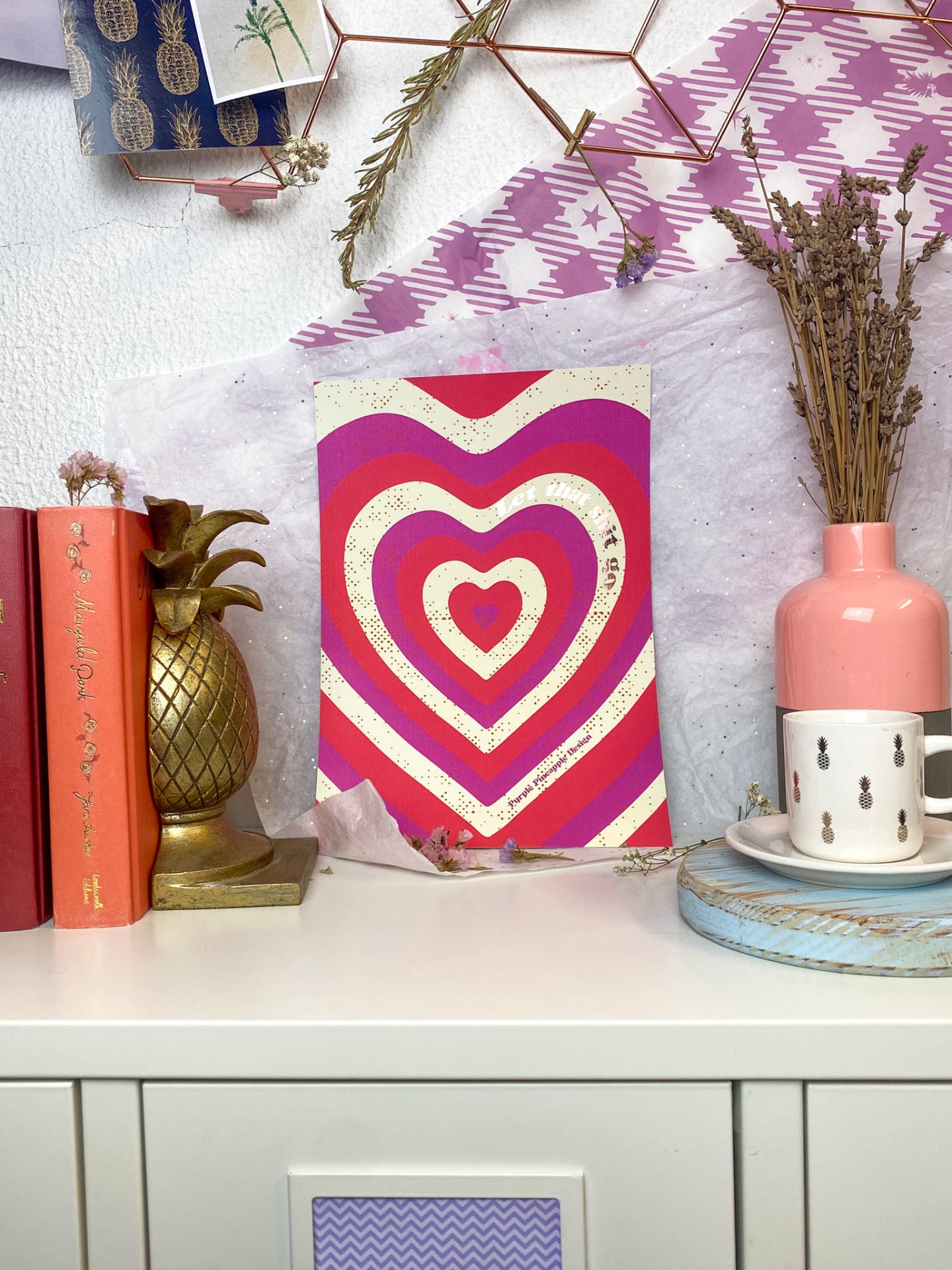 Decoração com quadro de corações coloridos, vaso rosa, caneca branca, suporte dourado de abacaxi e livros sobre superfície branca