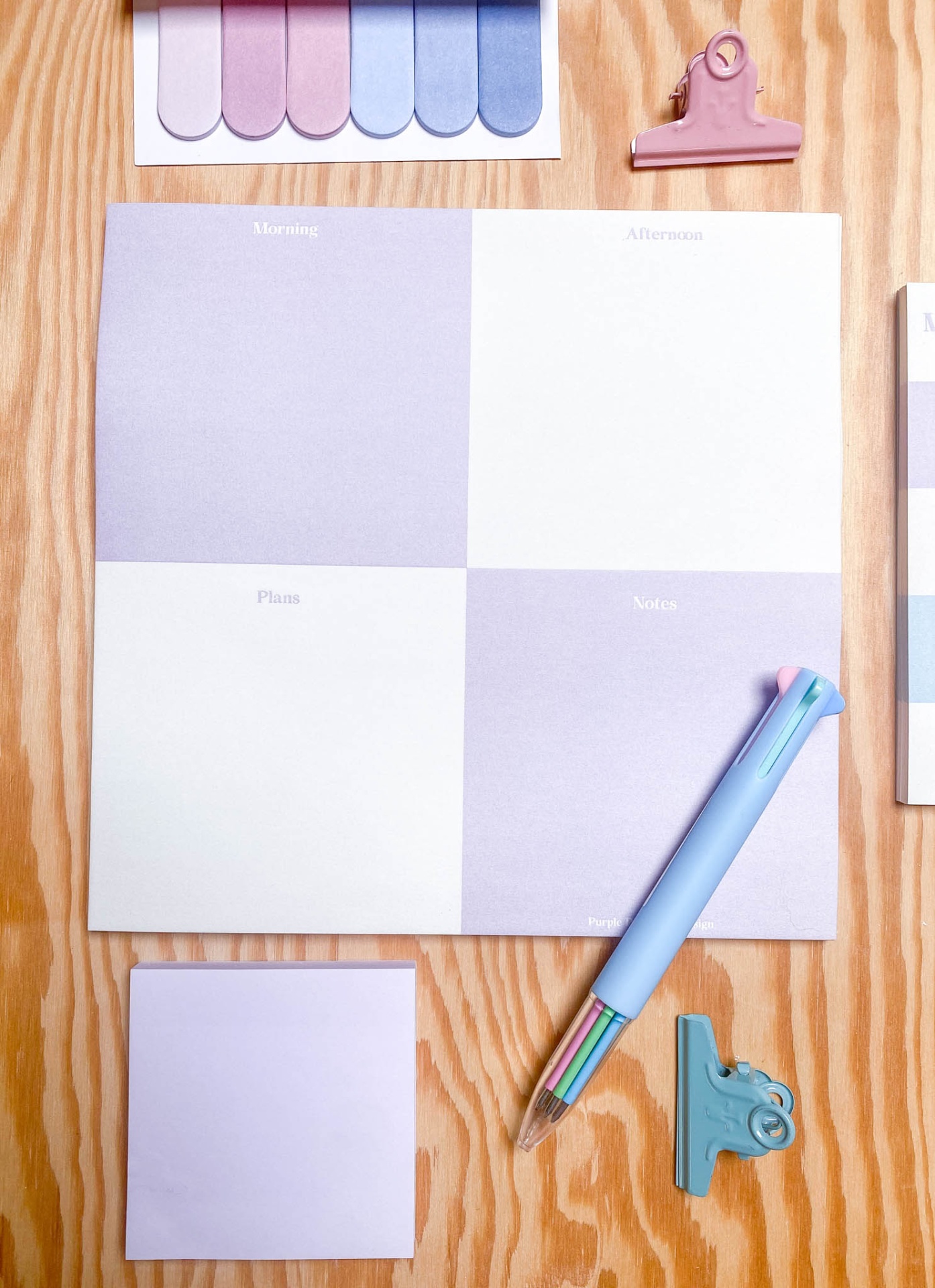 Material de escritório com bloco de notas quadrado lilás e branco, caneta colorida, clipes e amostras de cores sobre madeira clara