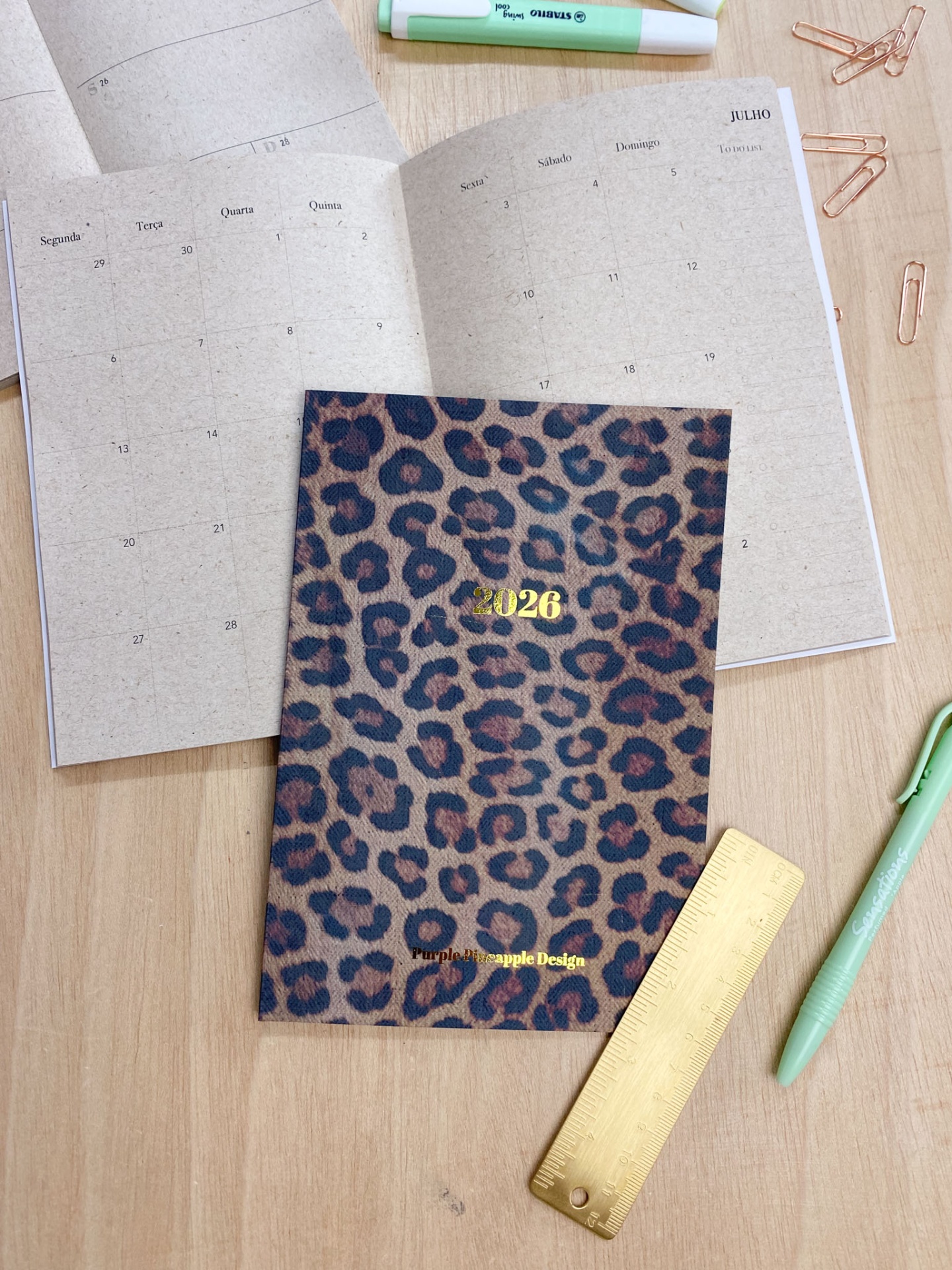 Agenda 2026 com capa estampada, papel de calendário aberto, régua dourada, caneta verde, clips em mesa de madeira.