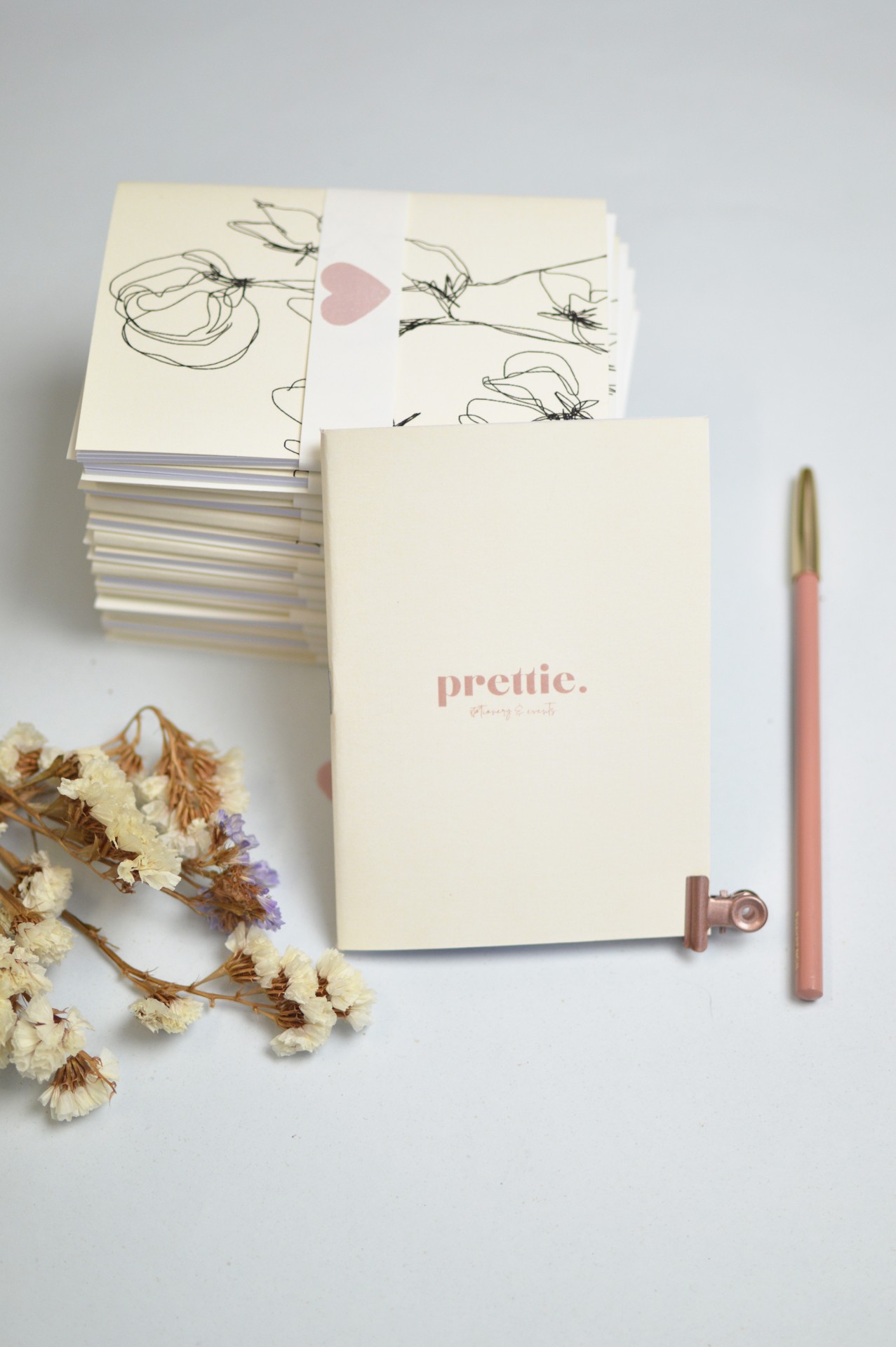 Caderno creme com texto 'prettie.', cartões florais, caneta rosa e folhas secas