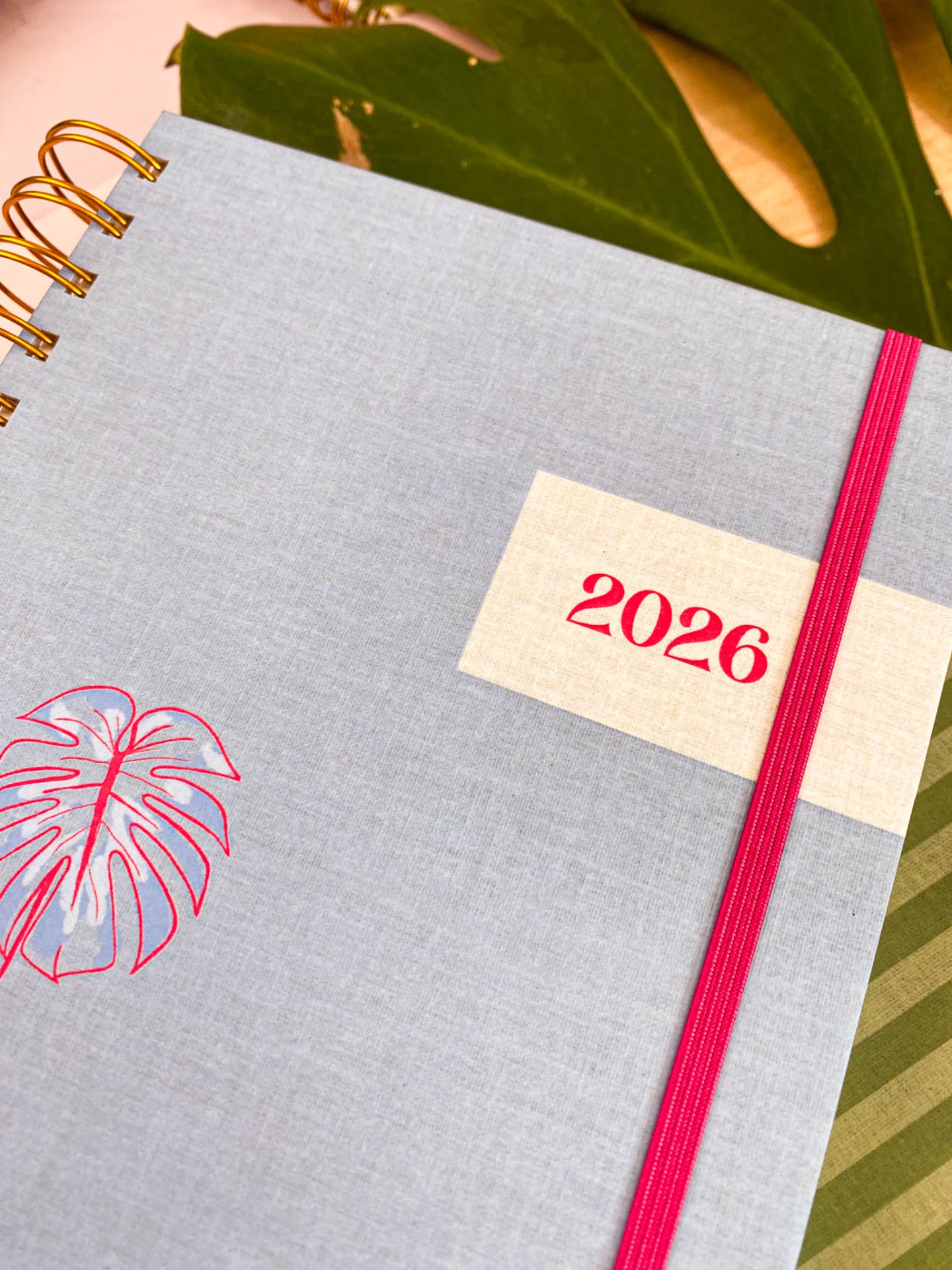 Agenda 2026 com capa cinzenta e detalhe rosa sobre folhas verdes